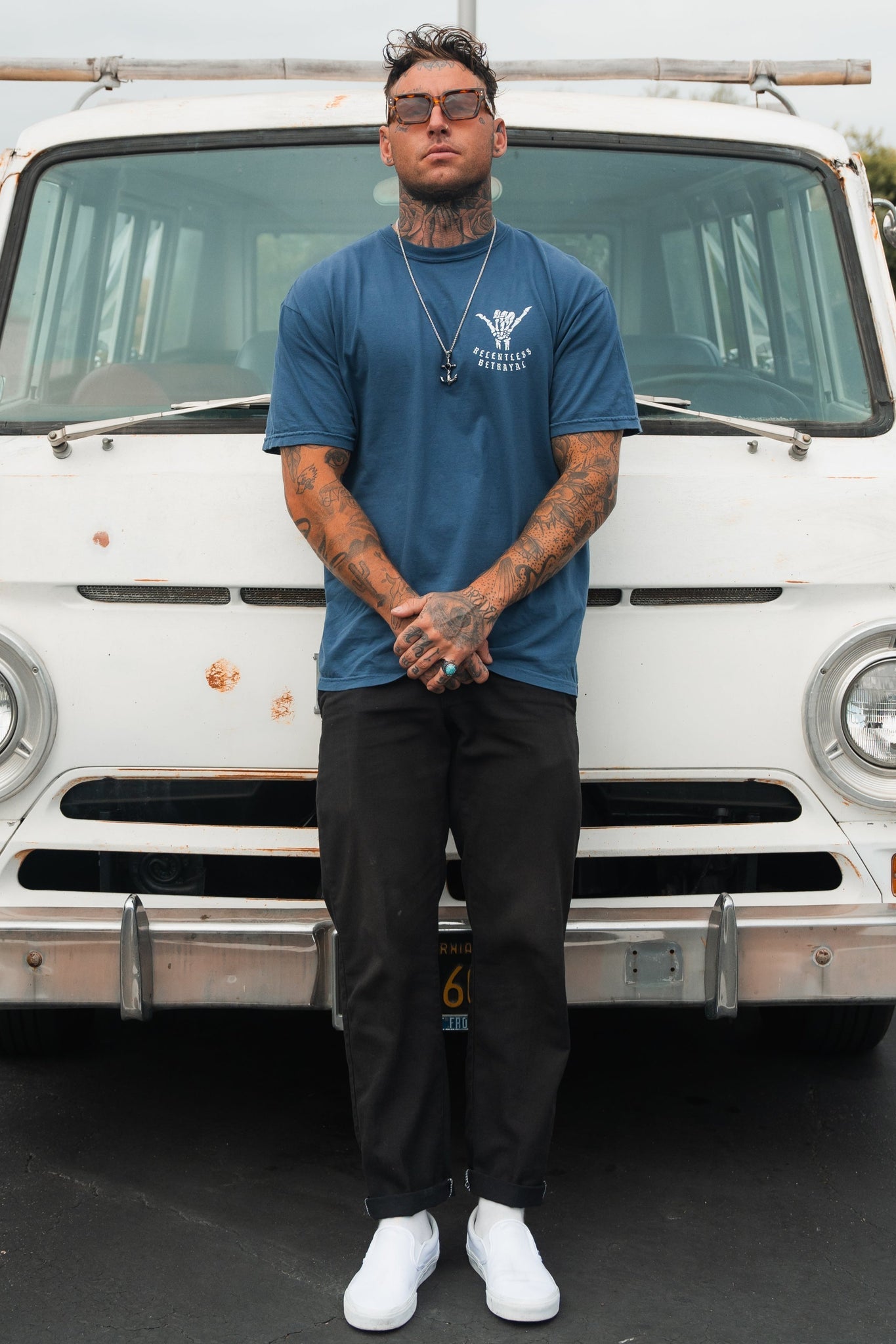 A tattooed man radiates resilience, confidently posing before a vintage white van. He wears sunglasses, black pants, white sneakers, and a Relentless Betrayals Sink or Swim Premium Slate T-Shirt. With a necklace adorning his neck and hands clasped in front, his stance echoes inner strength.