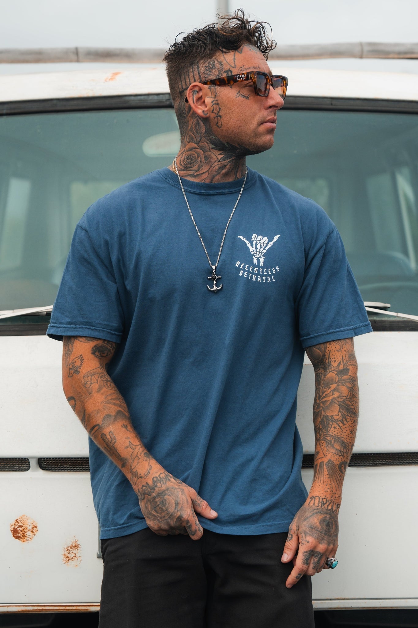 A tattooed man in a Relentless Betrayal Sink or Swim Premium Slate T-Shirt and dark pants stands before a rust-spotted, weathered white vehicle. His sunglasses, necklace, and confidently styled hair exude resilience. The vehicles rust symbolizes perseverance through time.