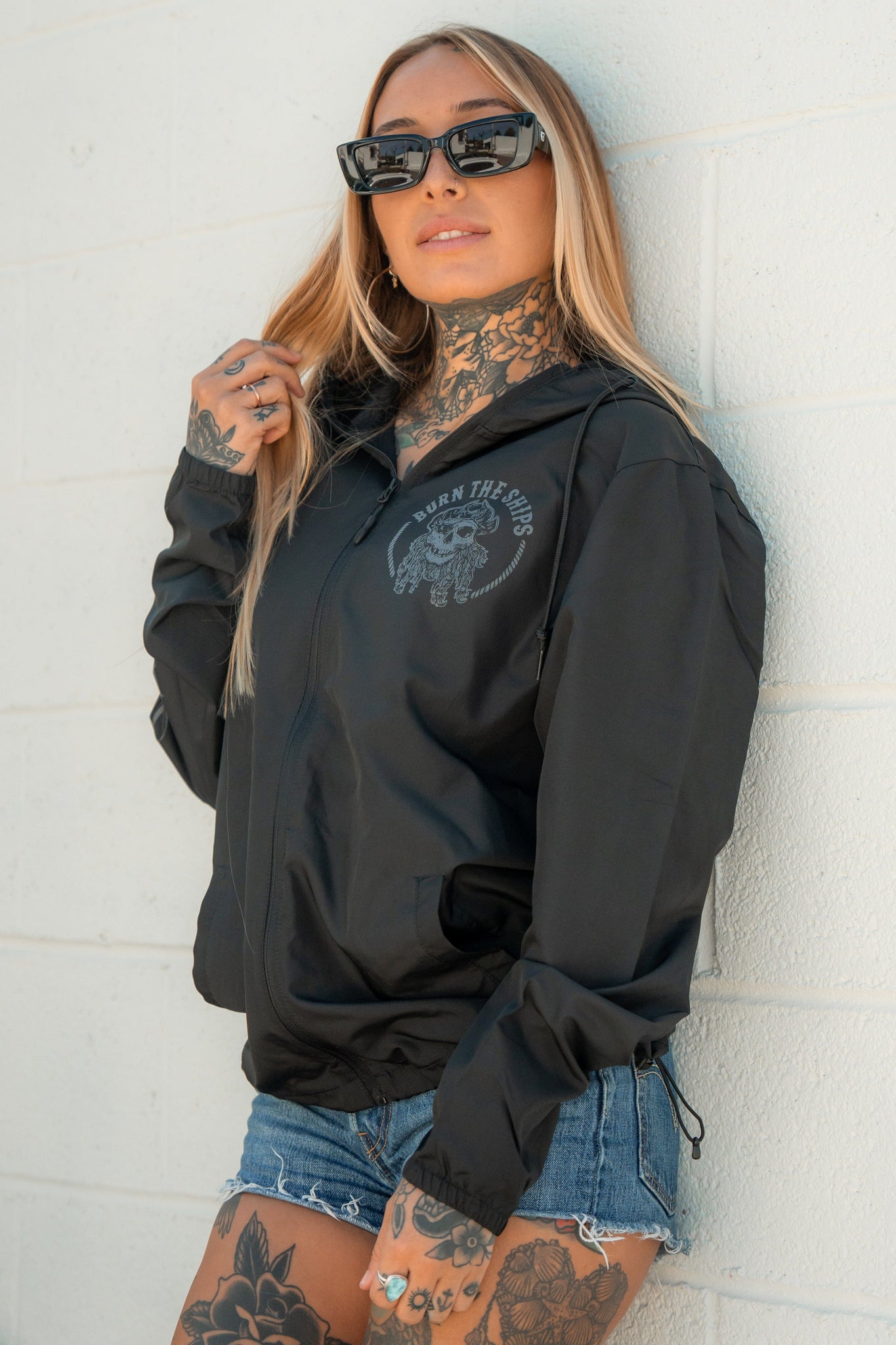 A person with long hair and tattoos wears sunglasses, denim shorts, and the Burn The Ships Windbreaker by Relentless Betrayal. They lean against a white wall with pirate lifestyle confidence, gazing directly at the camera.