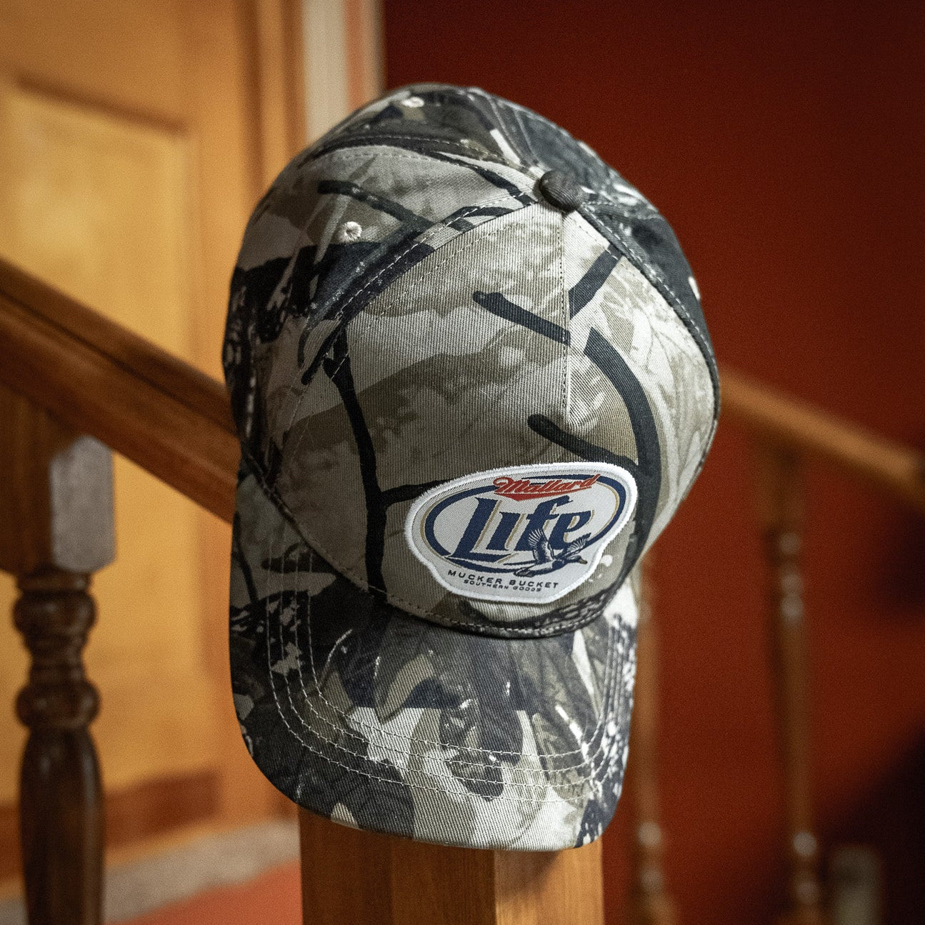 The Mallard Life Deadwood Camo Bucket by My Store is displayed on an indoor wooden staircase railing, with a blurred wooden door and red wall in the background.