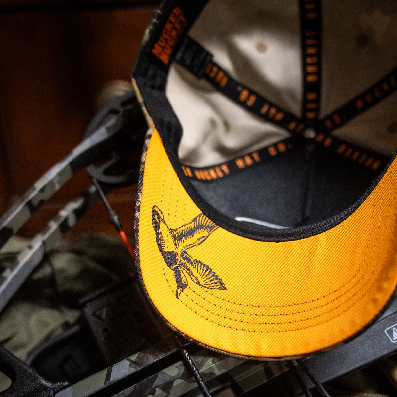 A close-up of the Mallard Life Deadwood Camo Bucket by My Store, featuring a bird illustration on the brim, resting on camo hunting gear.