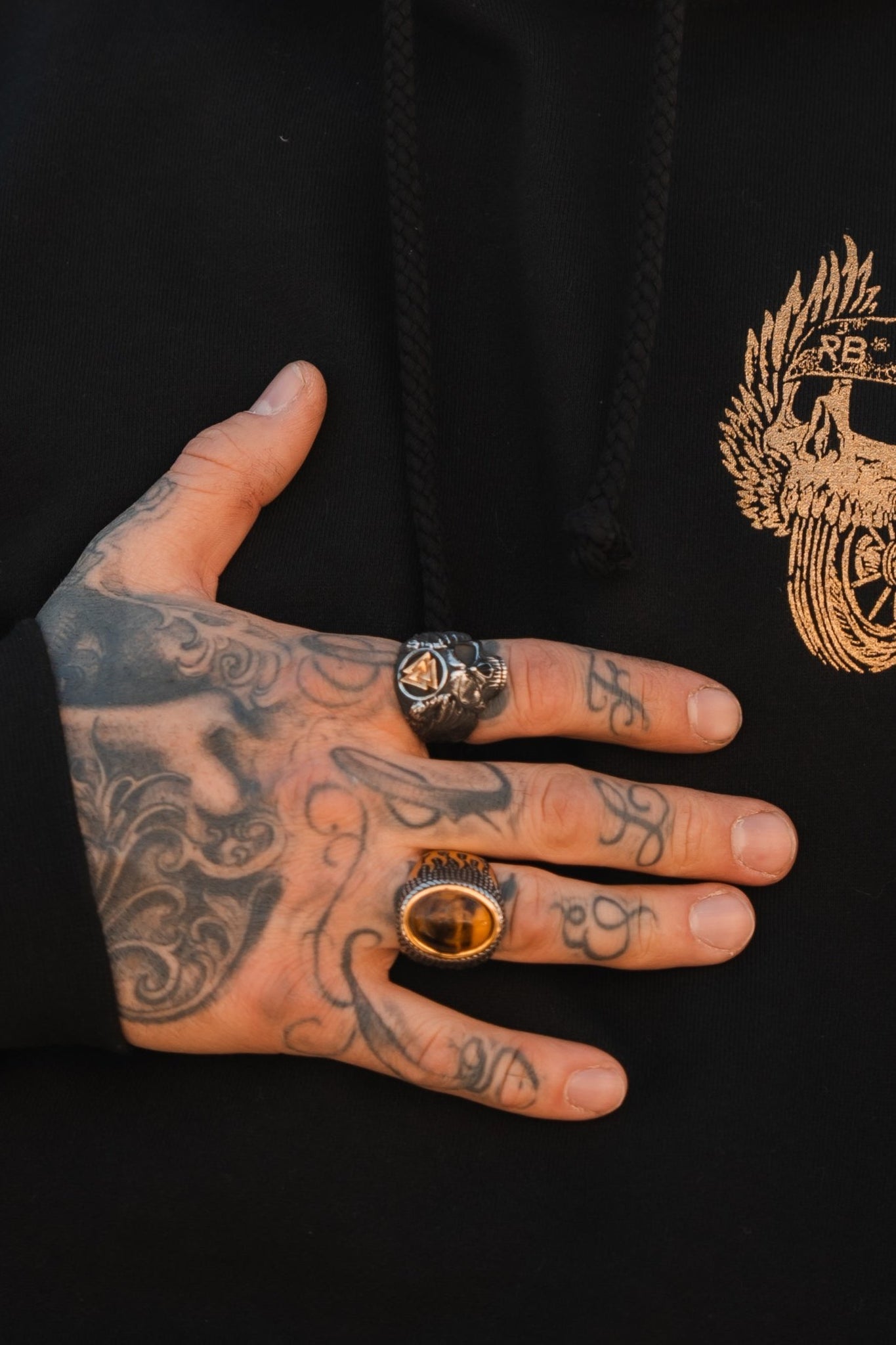 A tattooed hand on a black hoodie embodies youth culture with intricate designs, wearing Relentless Betrayals All Gas No Brakes Ring and another gold ring with a black stone. The hoodie, emphasizing individuality, features a gold embroidered chest emblem.