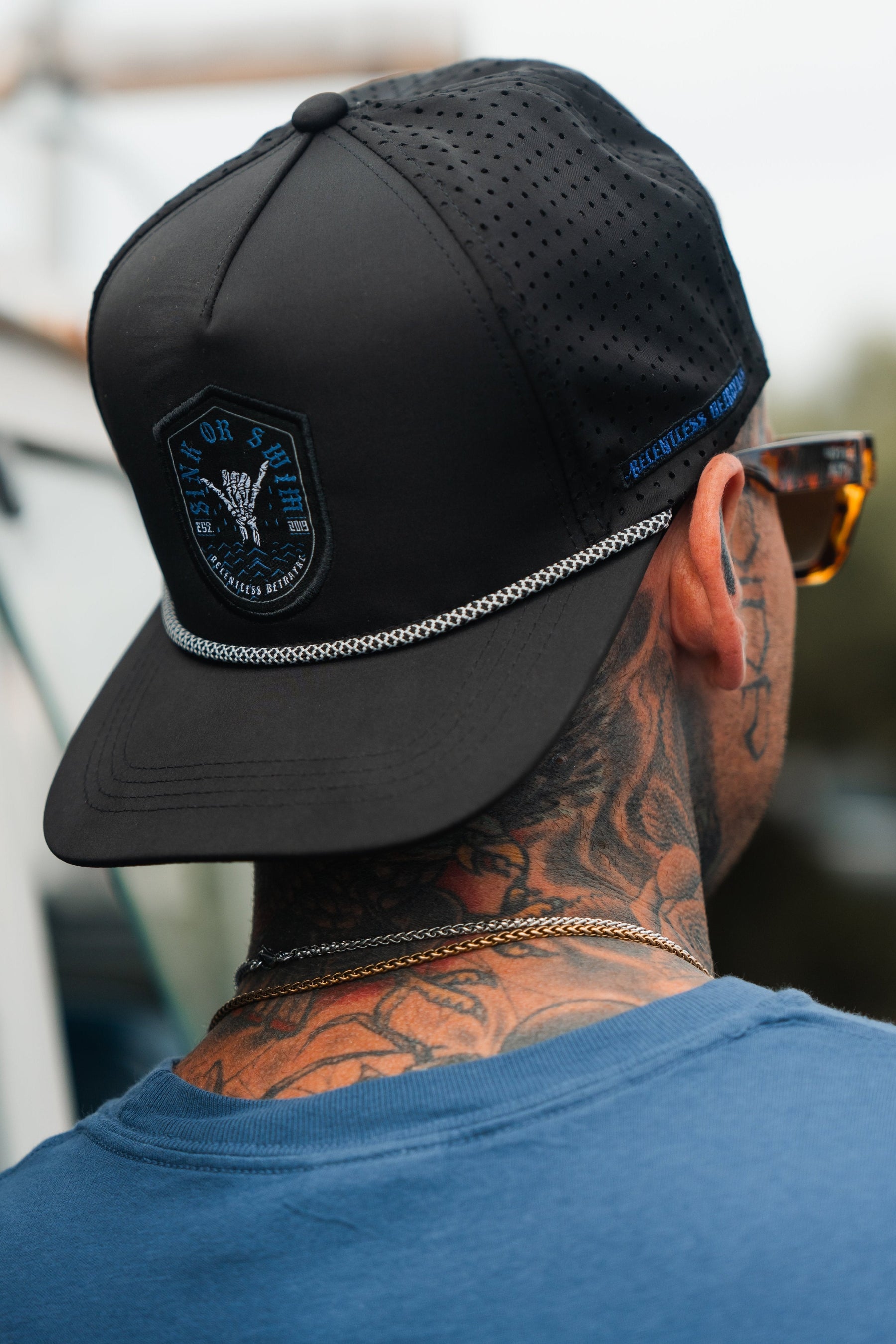 From the back, a tattooed individual wears a Relentless Betrayal Sink Or Swim Snapback in black, paired with a blue top. Sunglasses and a gold chain complete their look, embodying resilience against adversity.