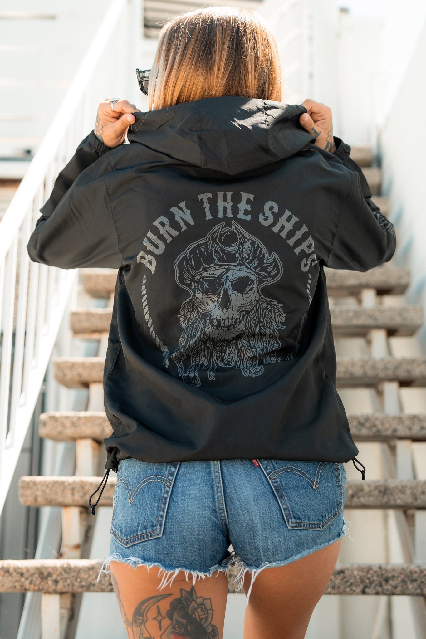 Embracing the pirate lifestyle, a person stands on outdoor stairs wearing the Burn The Ships Windbreaker by Relentless Betrayal. This black custom piece features a skull and BURN THE SHIPS on the back. They hold their hood, with ripped denim shorts and flowing hair completing the look.