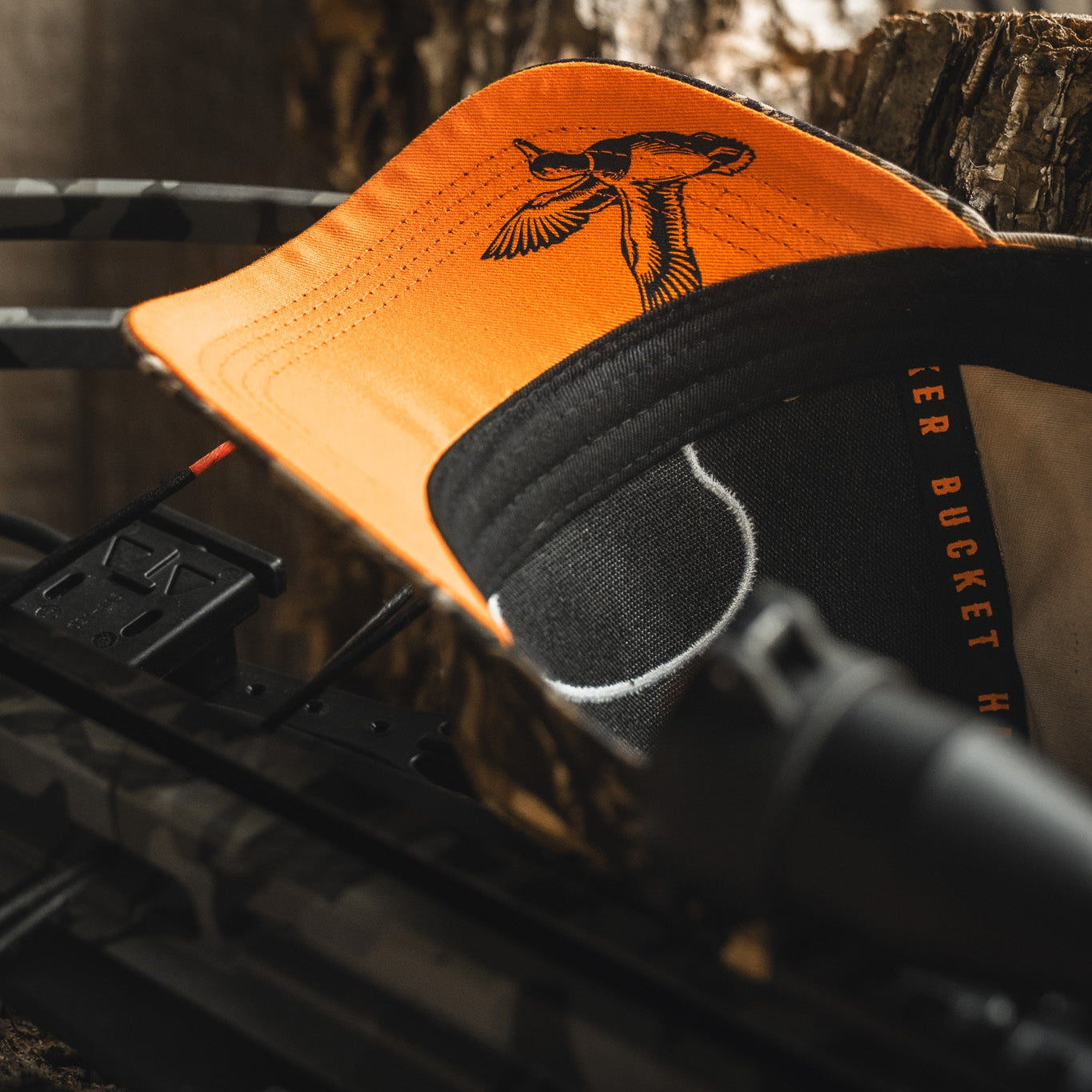 A close-up of the Mallard Life Deadwood Camo Bucket from My Store, featuring an orange and black design with a duck illustration under the brim, resting on a camo crossbow with a tree trunk in the background.