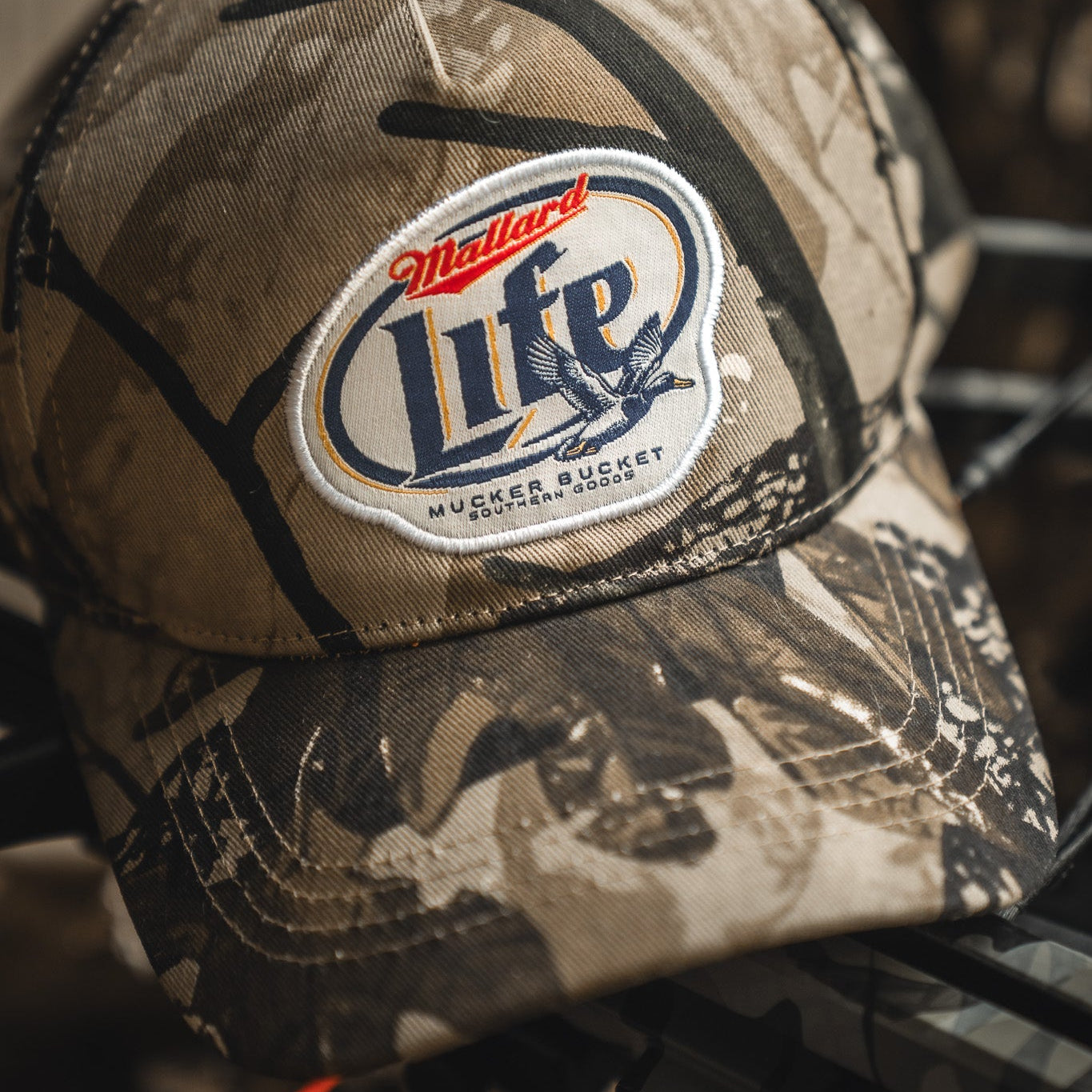 The Mallard Life Deadwood Camo Bucket by My Store is a camo snapback hat with a patch that reads "Mallard Life Mucker Bucket Southern Goods," including a flying duck graphic and brown-tan accents.