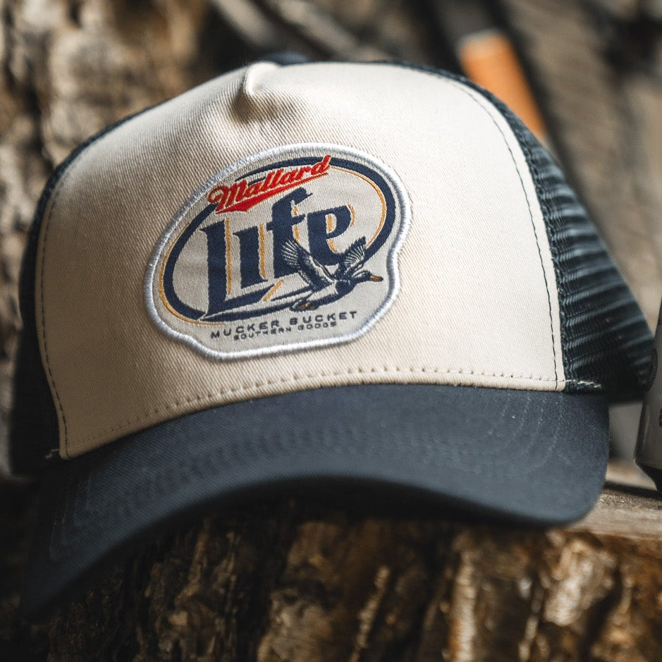 The Mallard Life Trucker Bucket by My Store, a black and white snapback with a detailed Mallard Life duck patch, sits on a wooden surface.