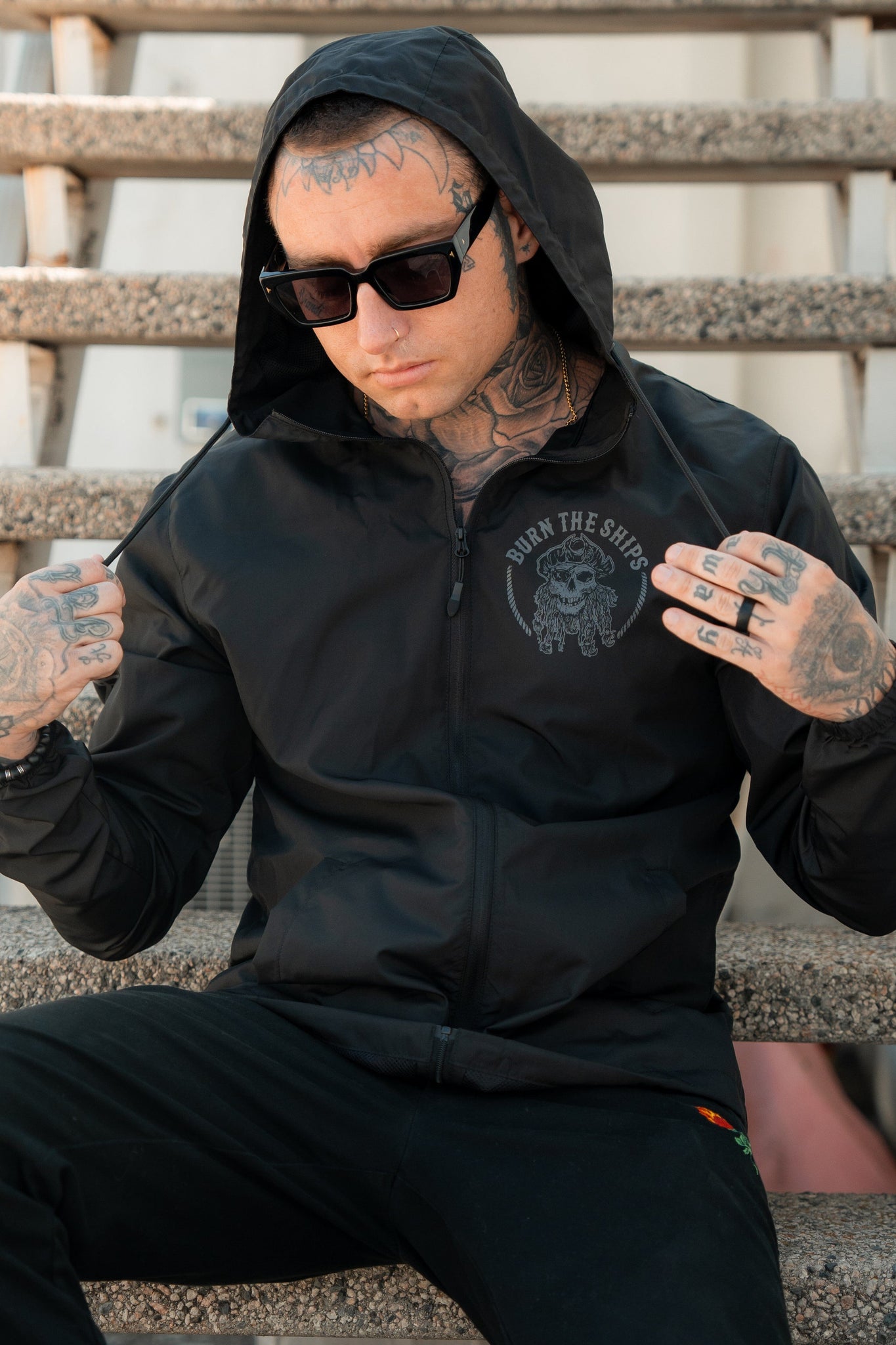 A person in a black Burn The Ships Windbreaker by Relentless Betrayal and sunglasses sits on stairs. Tattoos cover their face, neck, and hands. The jackets modern pirate lifestyle vibe stands out with its custom design and water-resistant practicality.