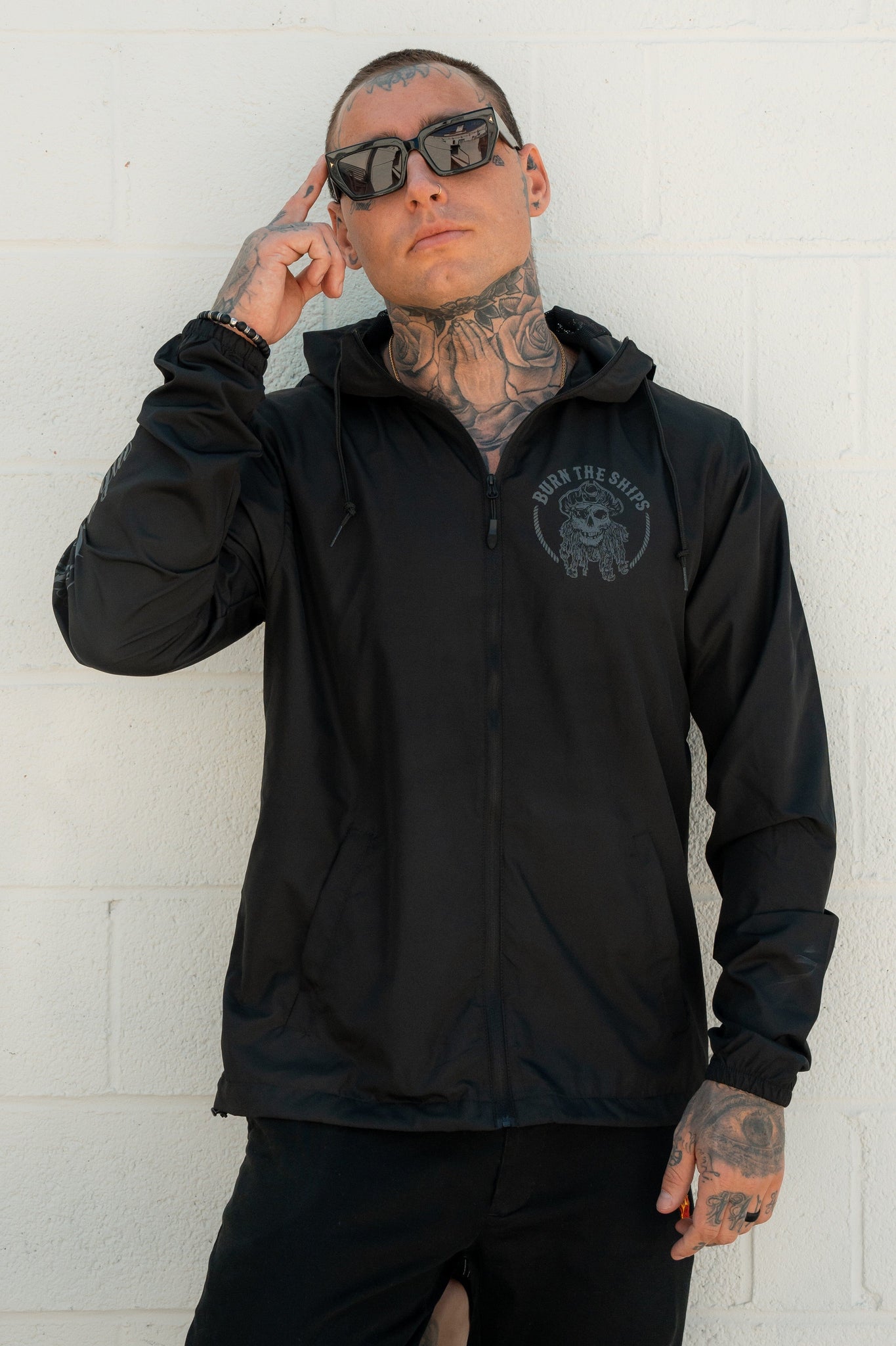 Dressed in a Burn The Ships Windbreaker by Relentless Betrayal featuring a water-resistant coating and dark sunglasses, a tattooed person stands confidently against a white brick wall, touching their temple with one hand, exuding a pirate-inspired vibe.