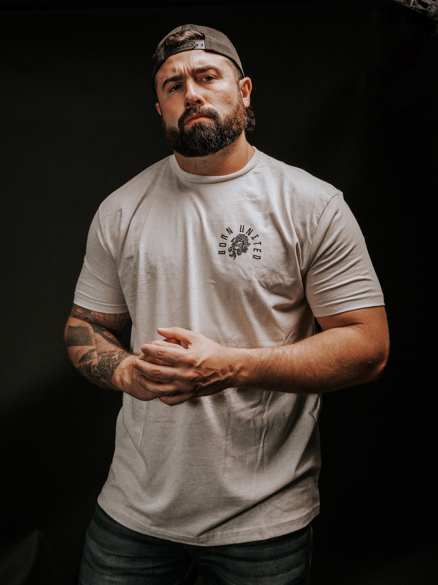 A bearded, tattooed man stands against a dark background, wearing a light gray Beware T-shirt by Born United Private Label and a backward baseball cap. With clasped hands and a focused look, he exudes mystery, as if discerning hidden truths beyond false facades.