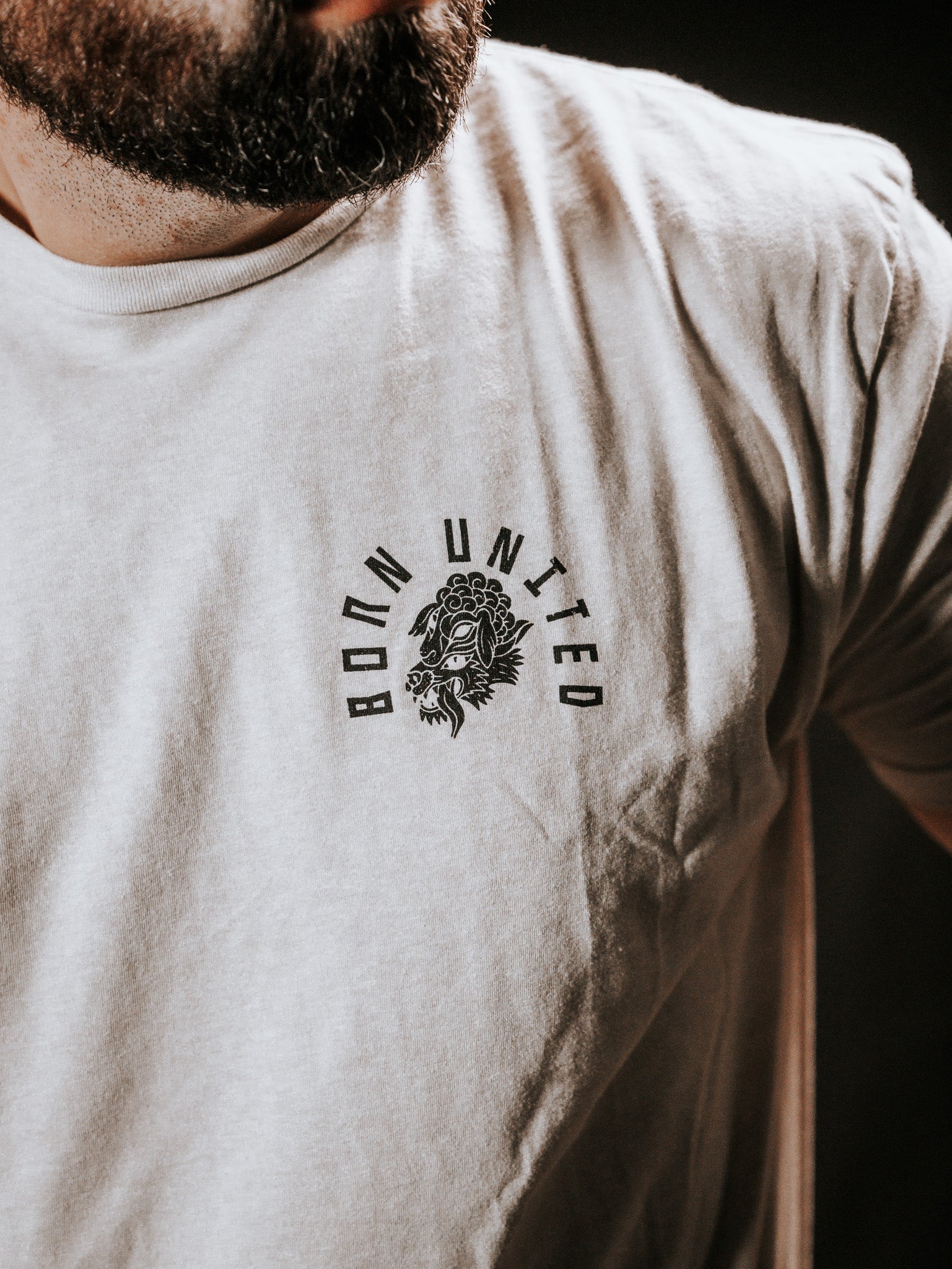 Close-up of someone wearing a white Born United Private Label Beware T-shirt, featuring BORN UNITED and a lion graphic on the chest against a dark background. The bold design stands out, warding off false prophets in sheeps clothing.