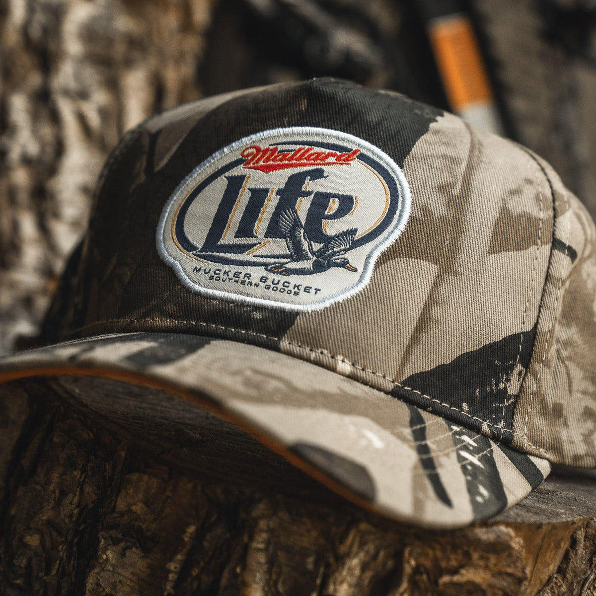 A Mallard Life Deadwood Camo Bucket hat by My Store sits on a tree trunk, showcasing a patch with Mallard Life and a flying duck.