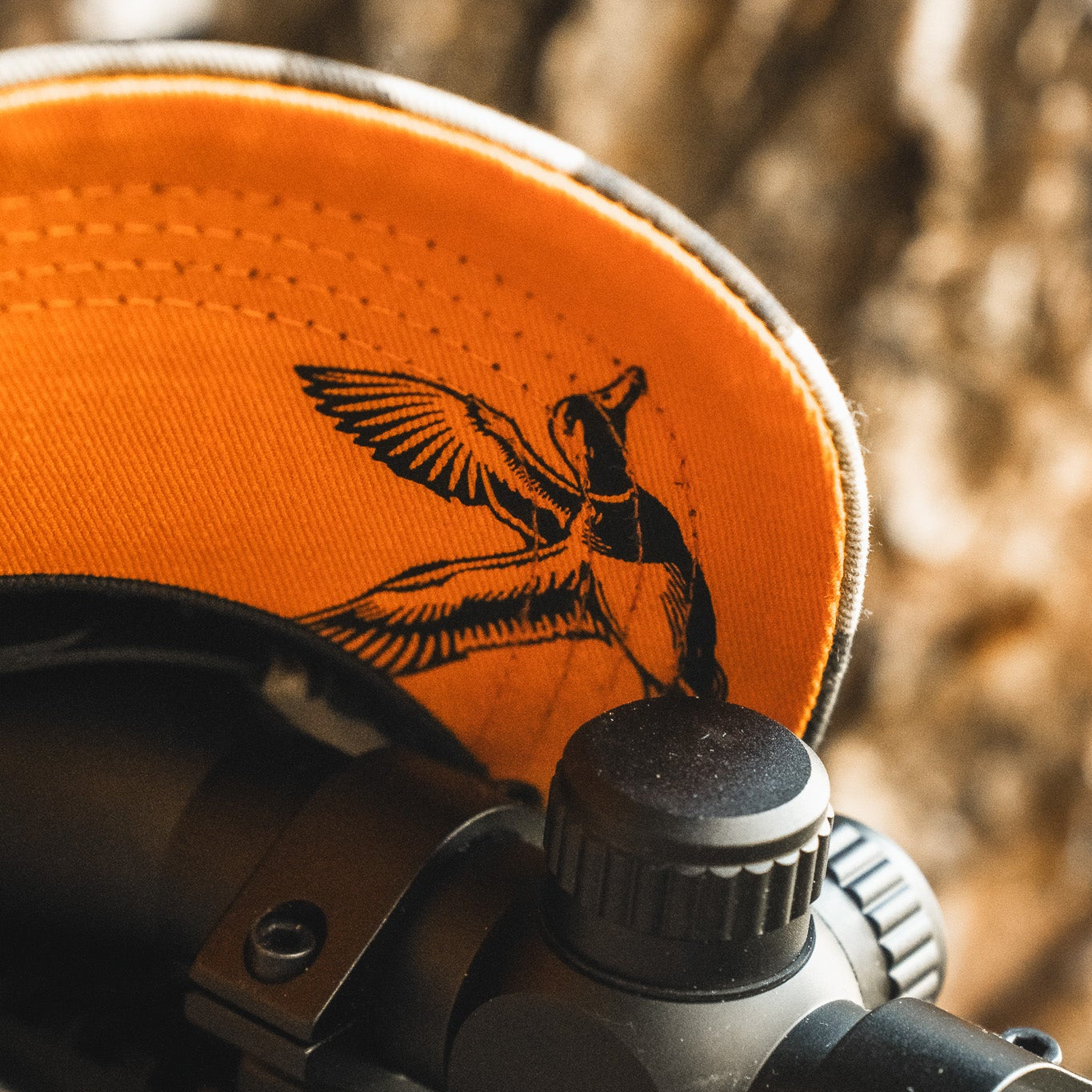 A close-up of the My Store Mallard Life Deadwood Camo Bucket hat with orange camo and a black bird print under the brim, resting on black metal equipment with dials, against a blurred brown background.