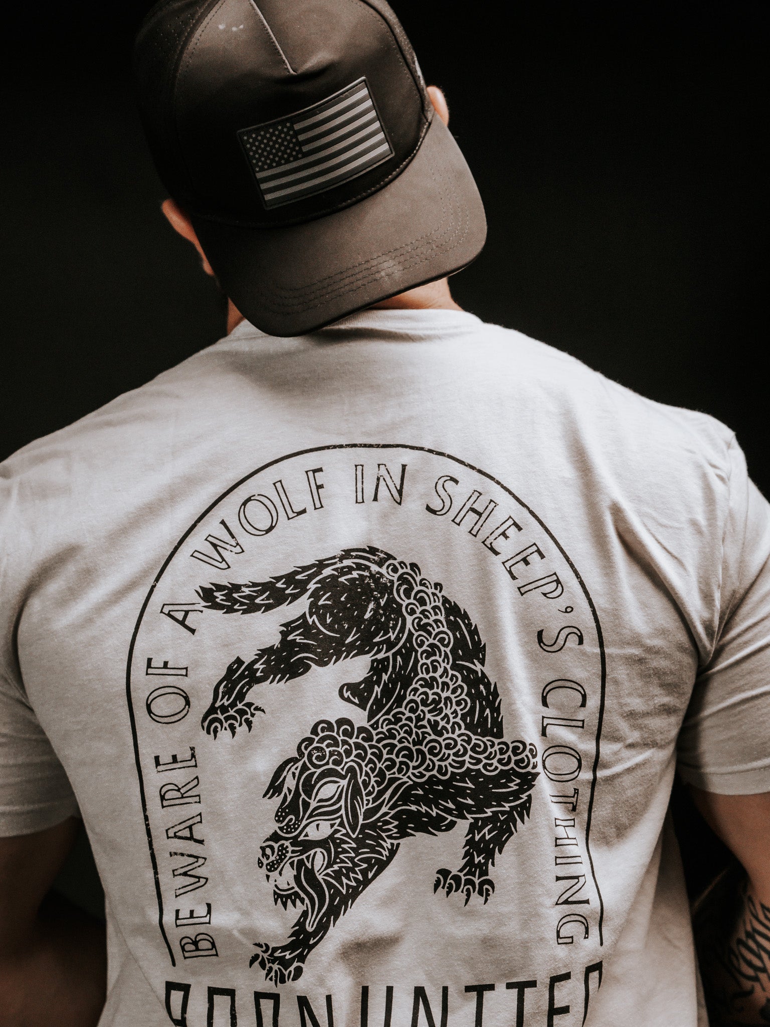 A person wearing a black cap with an American flag and a gray Beware shirt by Born United Private Label, featuring a wolf illustration, stands facing away. The shirt warns against false prophets with the phrase Beware of a Wolf in Sheeps Clothing.