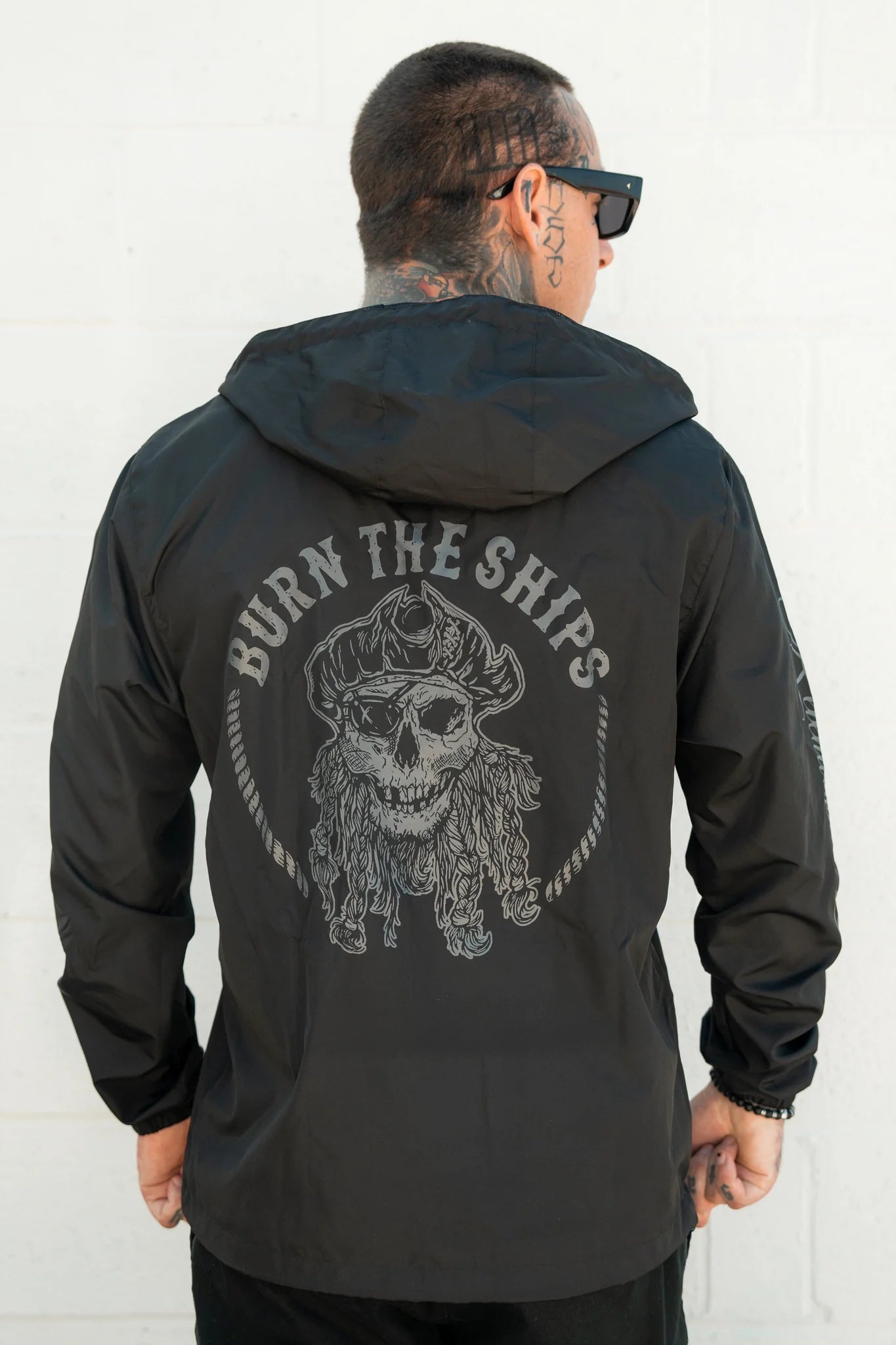 Against a light wall, the individual exudes confidence in the Burn The Ships Windbreaker from Relentless Betrayal. This custom black hooded jacket, adorned with a bold skull graphic and BURN THE SHIPS on the back, pairs perfectly with tattoos and sleek sunglasses to embody the pirate lifestyle.