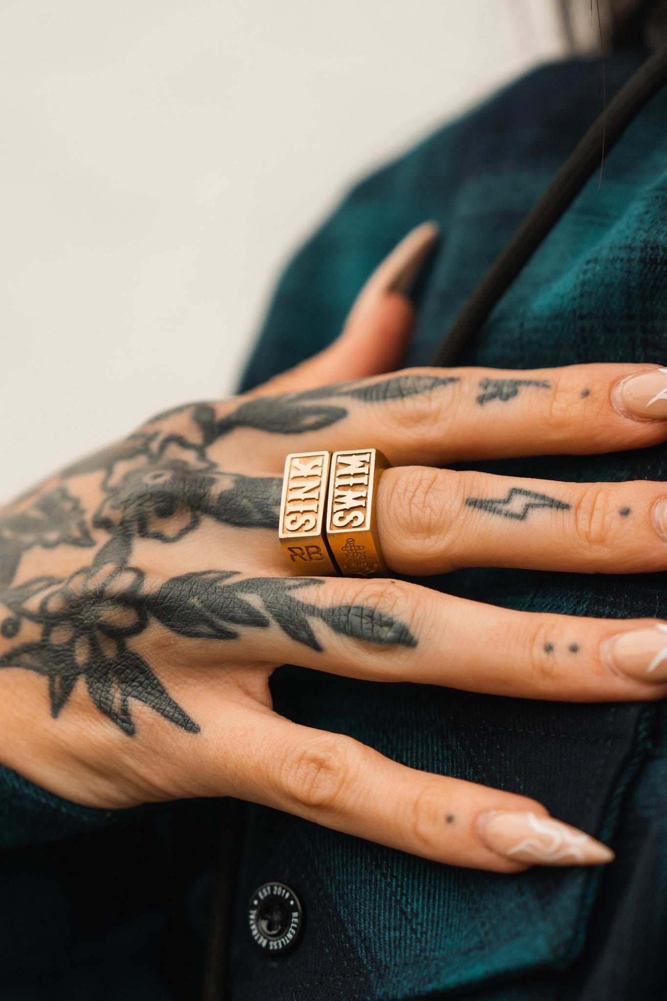 A close-up of a persons hand displays intricate floral tattoos and two Gold Sink or Swim Rings by Relentless Betrayal. The person, in a dark green plaid shirt, shows off pointed manicured nails. A handcrafted stainless steel ring enhances this striking ensemble.