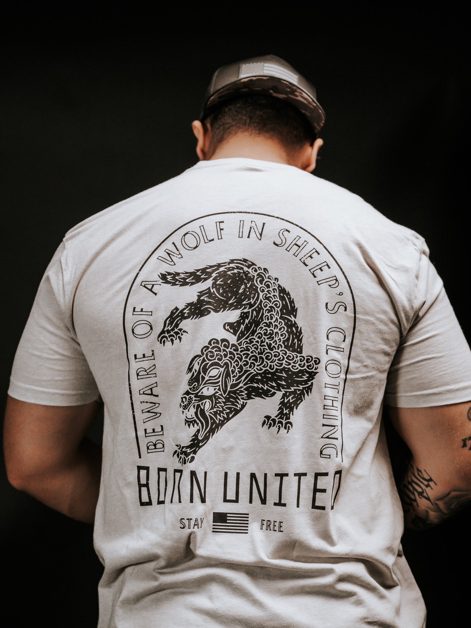 A person wears a light-colored t-shirt with a snarling wolf illustration and the words Beware of ravening wolves in sheeps clothing, plus Born United, Stay Free, and a small American flag. The brand is Born United Private Label, and the product name is Beware.