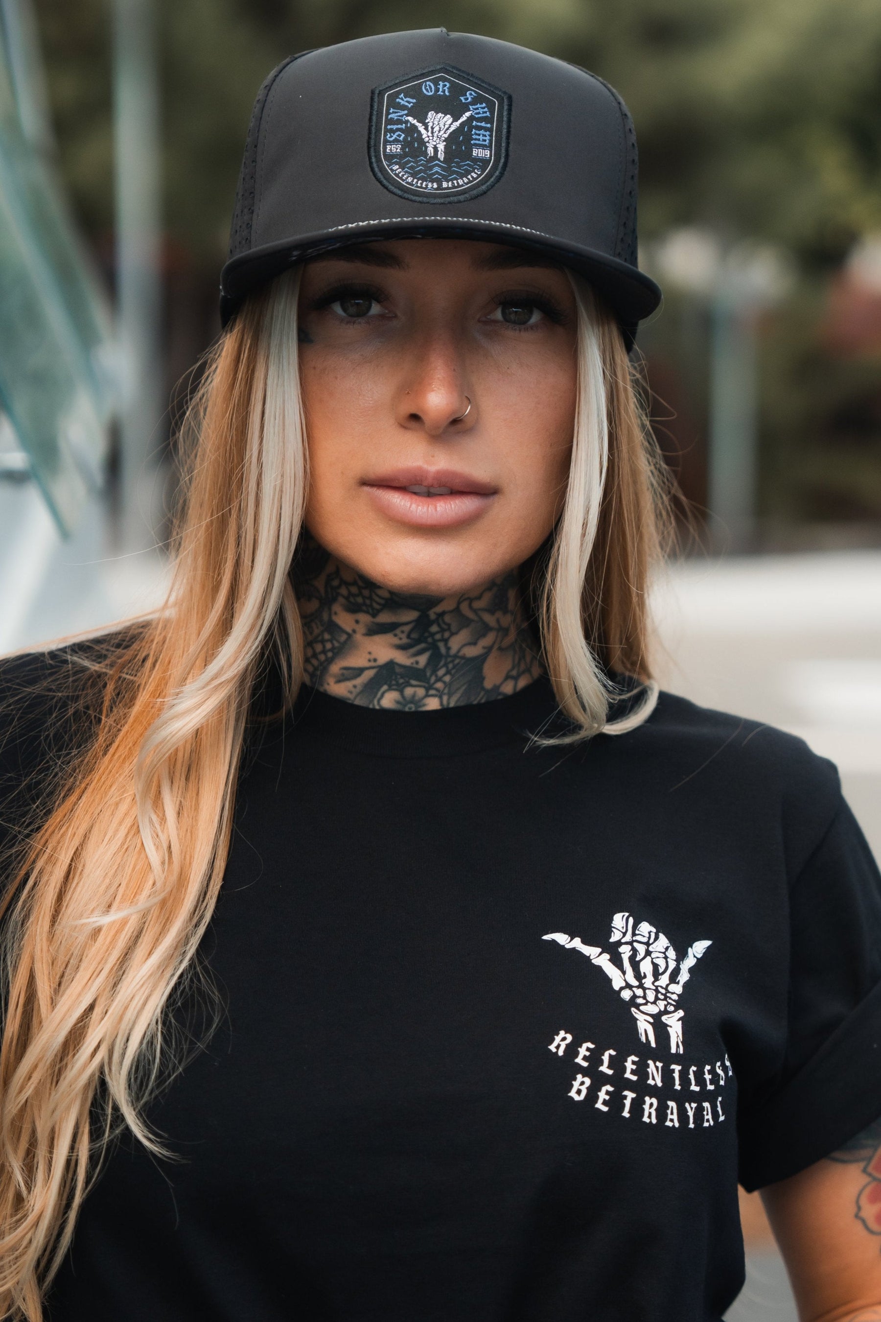 A person with long blonde hair, wearing a Relentless Betrayal Sink Or Swim Snapback and t-shirt, stands outdoors radiating resilience. Tattoos snake up their neck as they face a blurred background of trees and a building, embodying quiet perseverance.