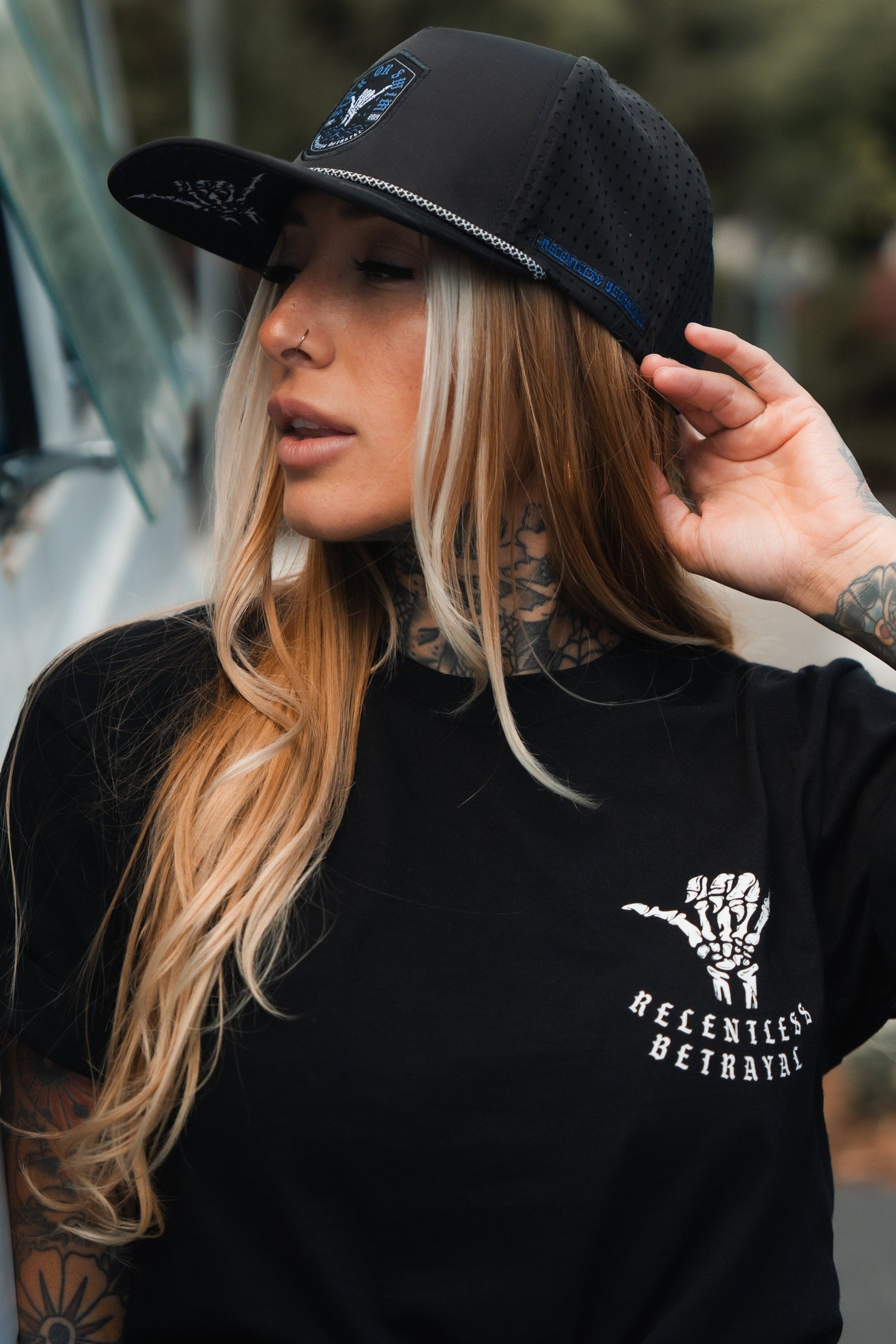 A long-haired, tattooed individual in a black Sink Or Swim Snapback by Relentless Betrayal and a resilience-themed black t-shirt touches their cap while looking aside, exuding an overcoming spirit against a blurred background.