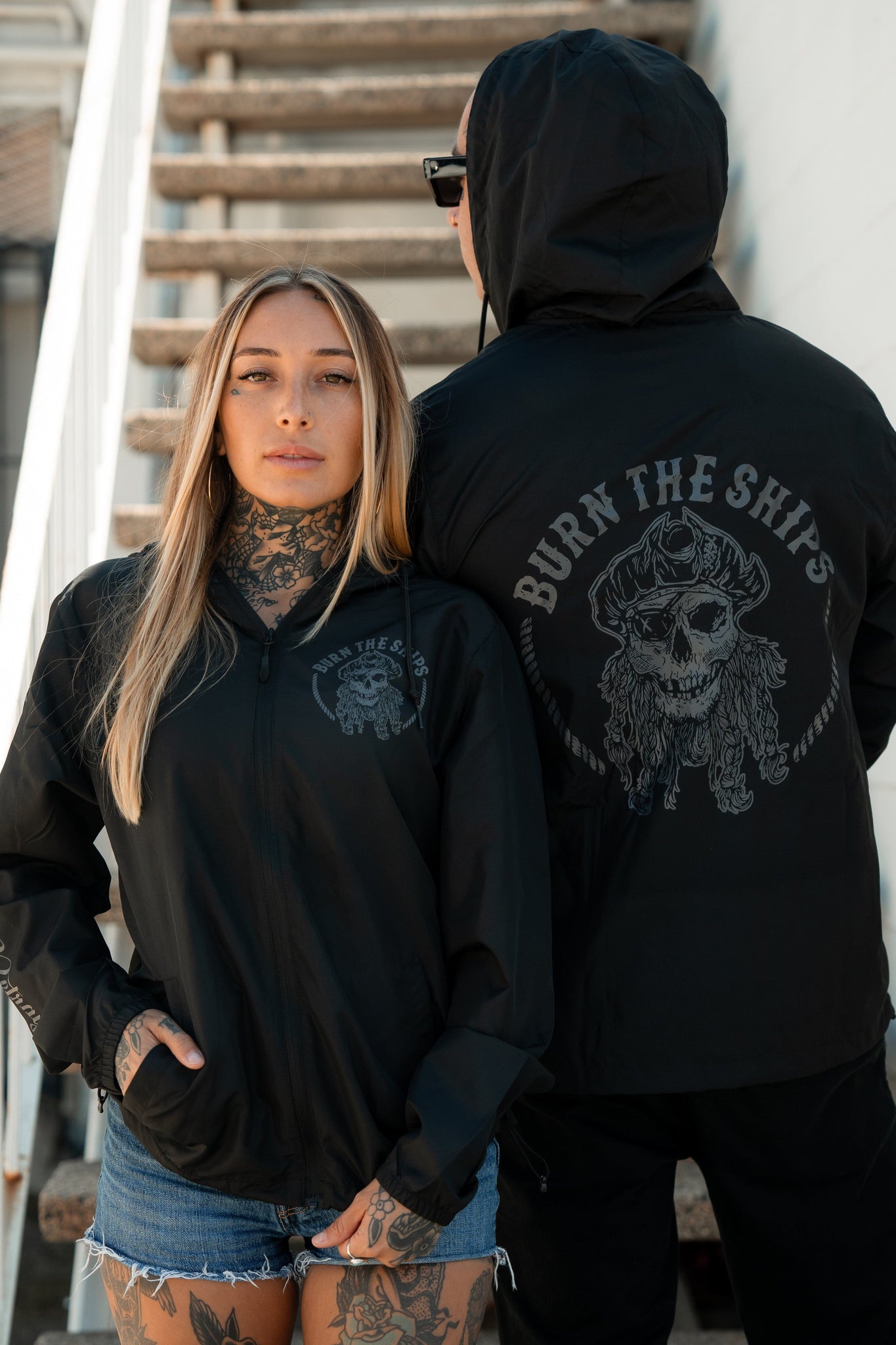 Two people on stairs wear Relentless Betrayals Burn The Ships Windbreakers, featuring a skull and crossbones design. The front person shows off long blonde hair and tattoos, while the back one wears a hood, channeling a pirate vibe.