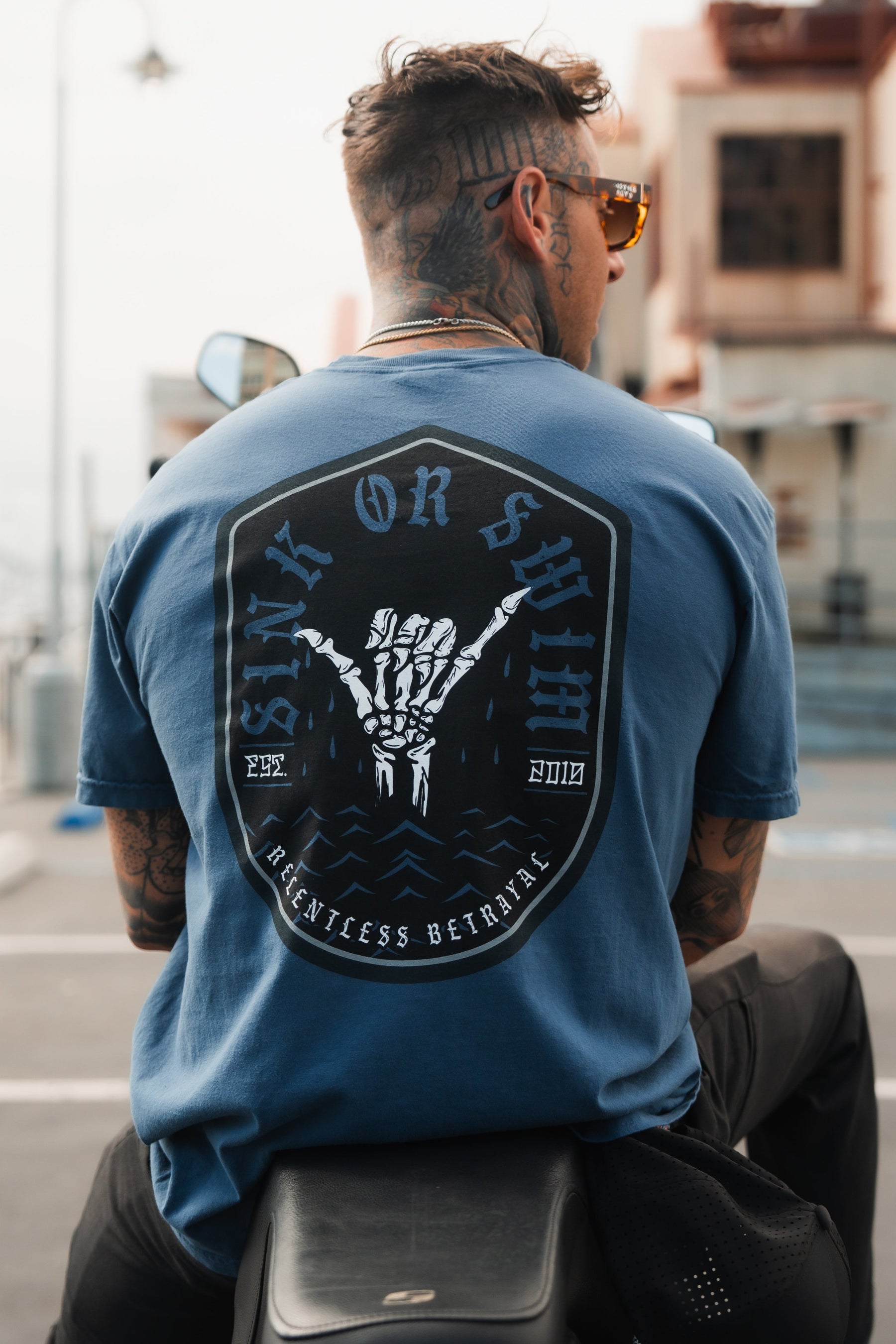 Wearing the Sink or Swim Premium Slate T-Shirt by Relentless Betrayal, with its bold tattooed arms design and powerful phrases, a person sits on a motorcycle. The scene captures resilience and inner strength on the open road.