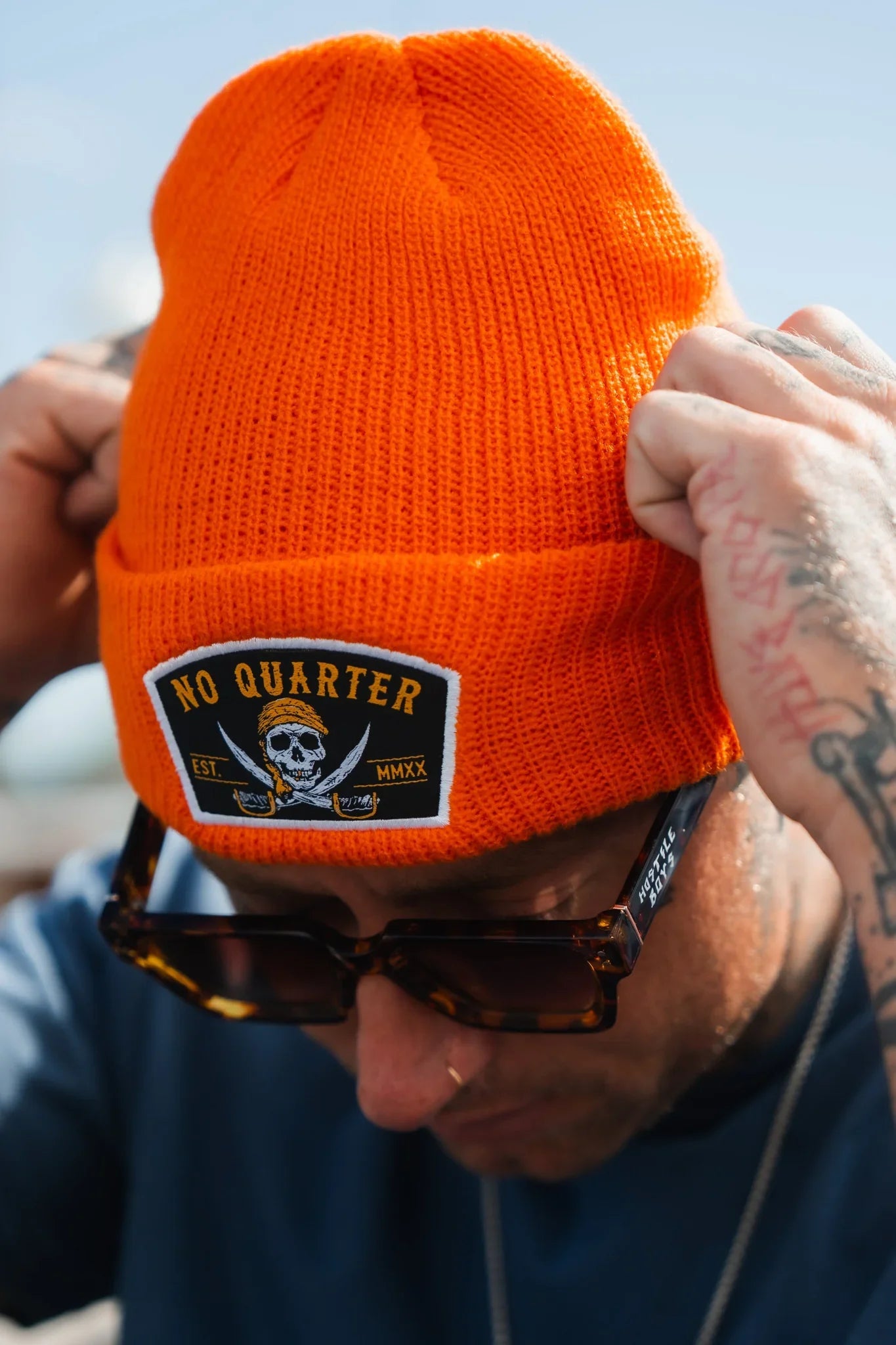 A person with tattooed arms adjusts their Relentless Betrayal No Quarter Statement Beanie, crafted from 100% acrylic and featuring a skull and crossed swords patch. The blurred background highlights the vivid orange hue and dark sunglasses.