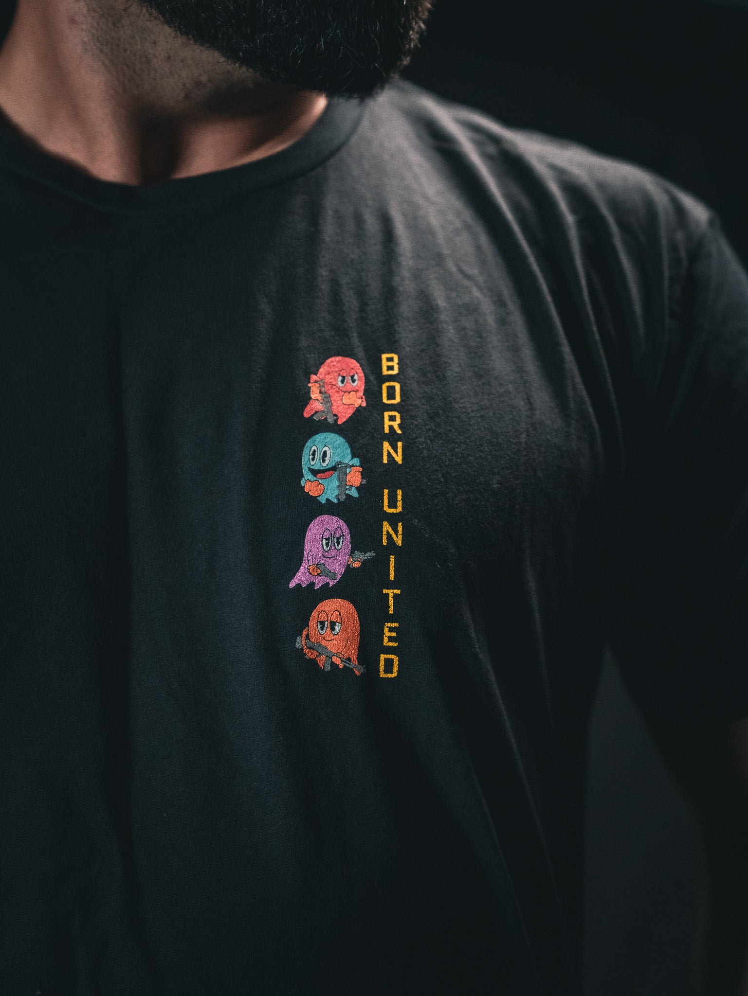 A person is wearing a Tac-Man (Warehouse) T-shirt from Born United Private Label. It features four vibrant cartoon ghosts stacked vertically on a dark backdrop, with BORN UNITED in yellow beside them.
