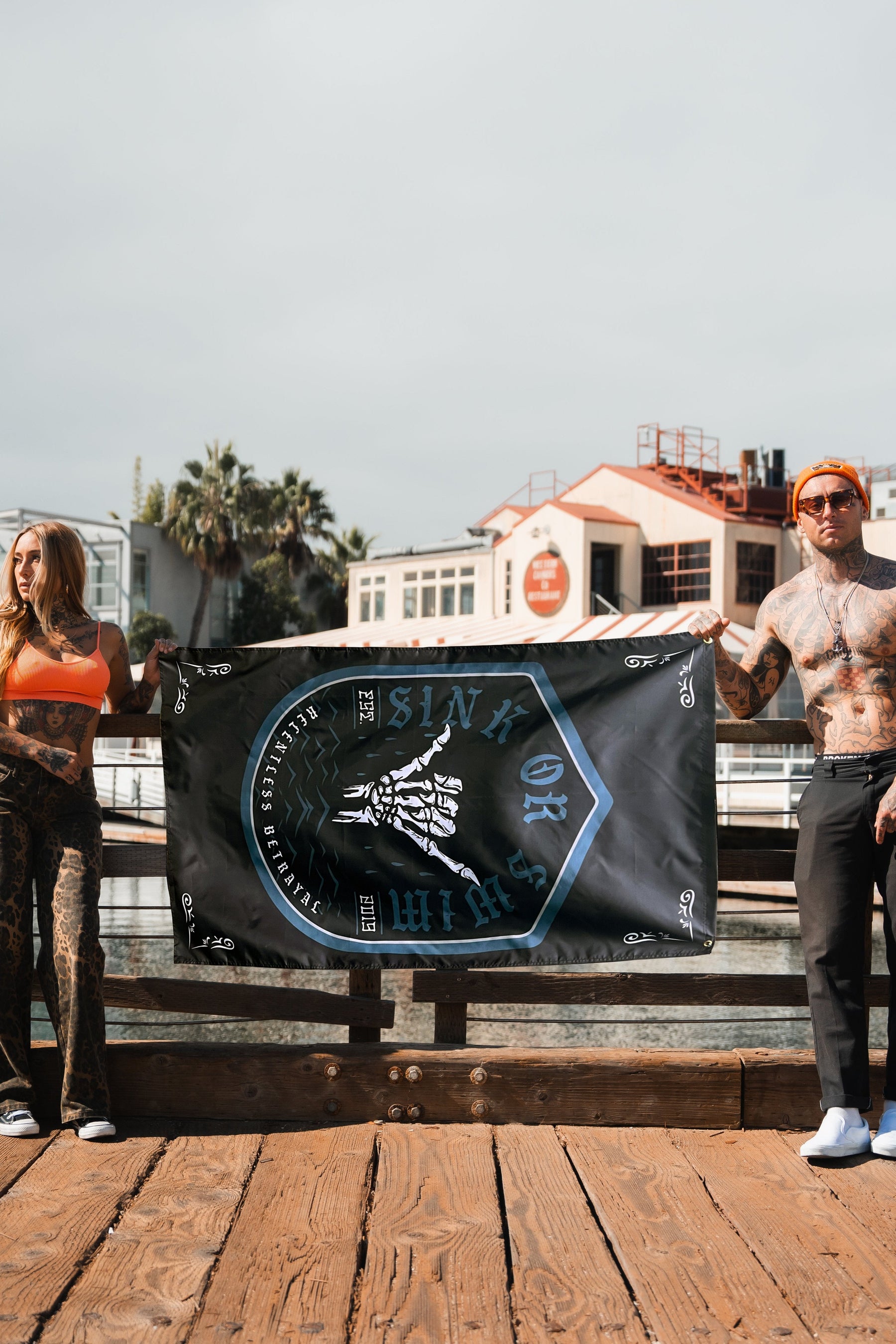 On a wooden dock, two people embody resilience as they hold the Relentless Betrayal Sink or Swim 2.O Flag, featuring blue and white designs on black. Buildings and palm trees rise in the background, symbolizing fortitude against the clear sky.