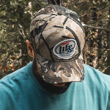 A bearded person in a teal shirt wears the My Store "Mallard Life Deadwood Camo Bucket" hat, looking down, with a blurred backdrop of trees and foliage.