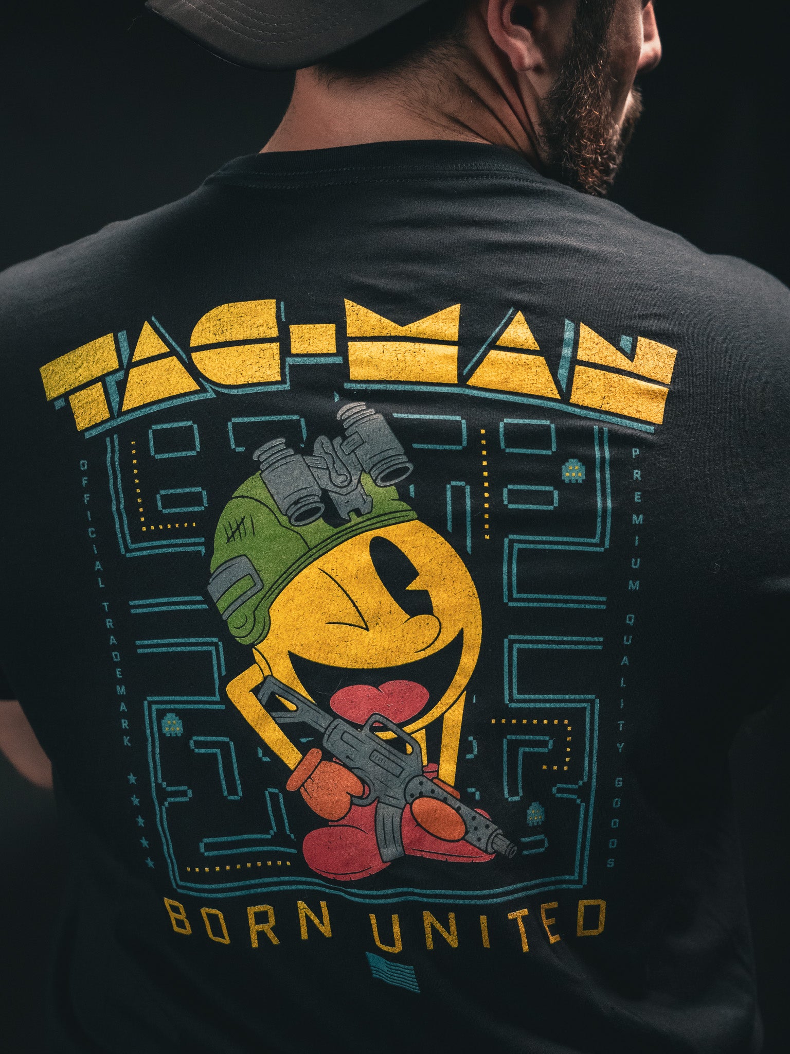 A man wears a black T-shirt featuring a yellow, round character in a helmet and boots, holding a gun. Tac-Man is above and Born United Private Label below the character, all in retro video game style.