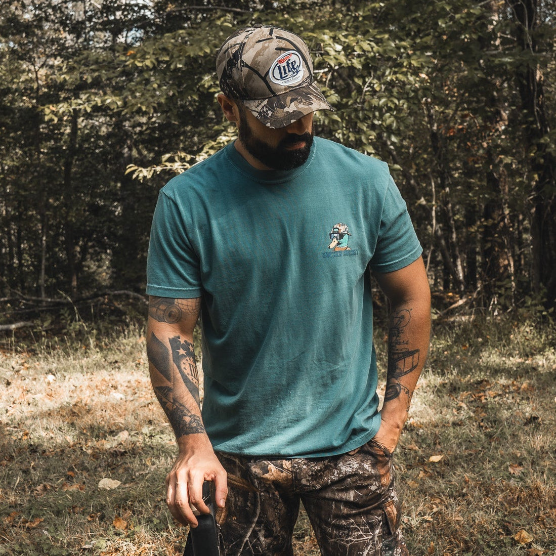 A bearded man with tattooed arms stands in a wooded area wearing the Mallard Life Deadwood Camo Bucket by My Store, along with a teal T-shirt and camouflage pants, holding a dark object and looking down amid the trees.