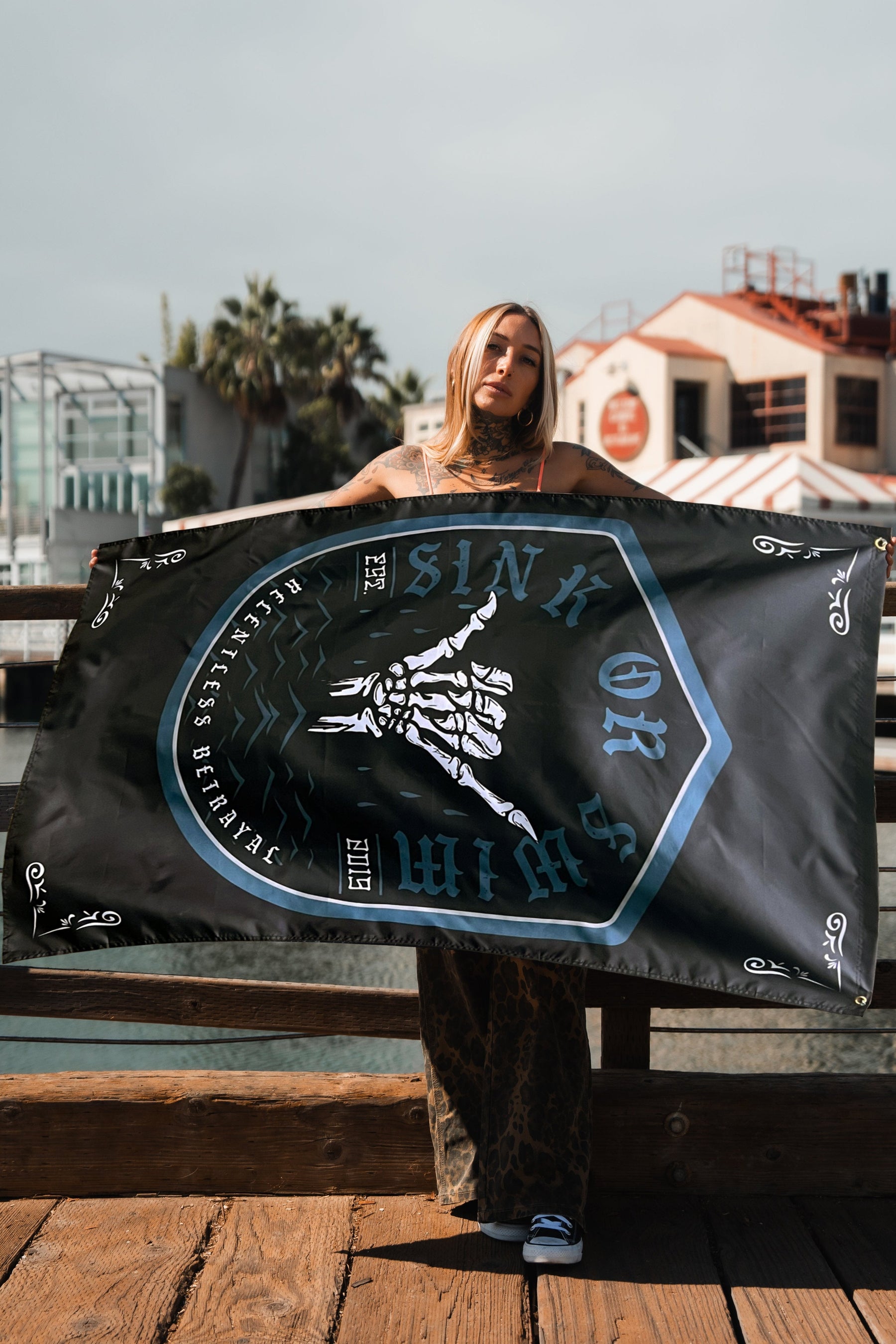 On a wooden dock, someone holds the Relentless Betrayal Sink or Swim 2.0 Flag, featuring a skeleton hand illustration and embodying resilience. In the background, buildings stand against a cloudy sky.