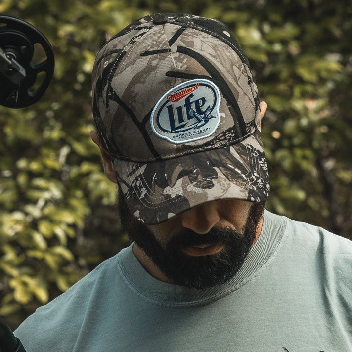 A bearded man in a Mallard Life Deadwood Camo Bucket hat by My Store looks down while standing outdoors with green foliage in the background.