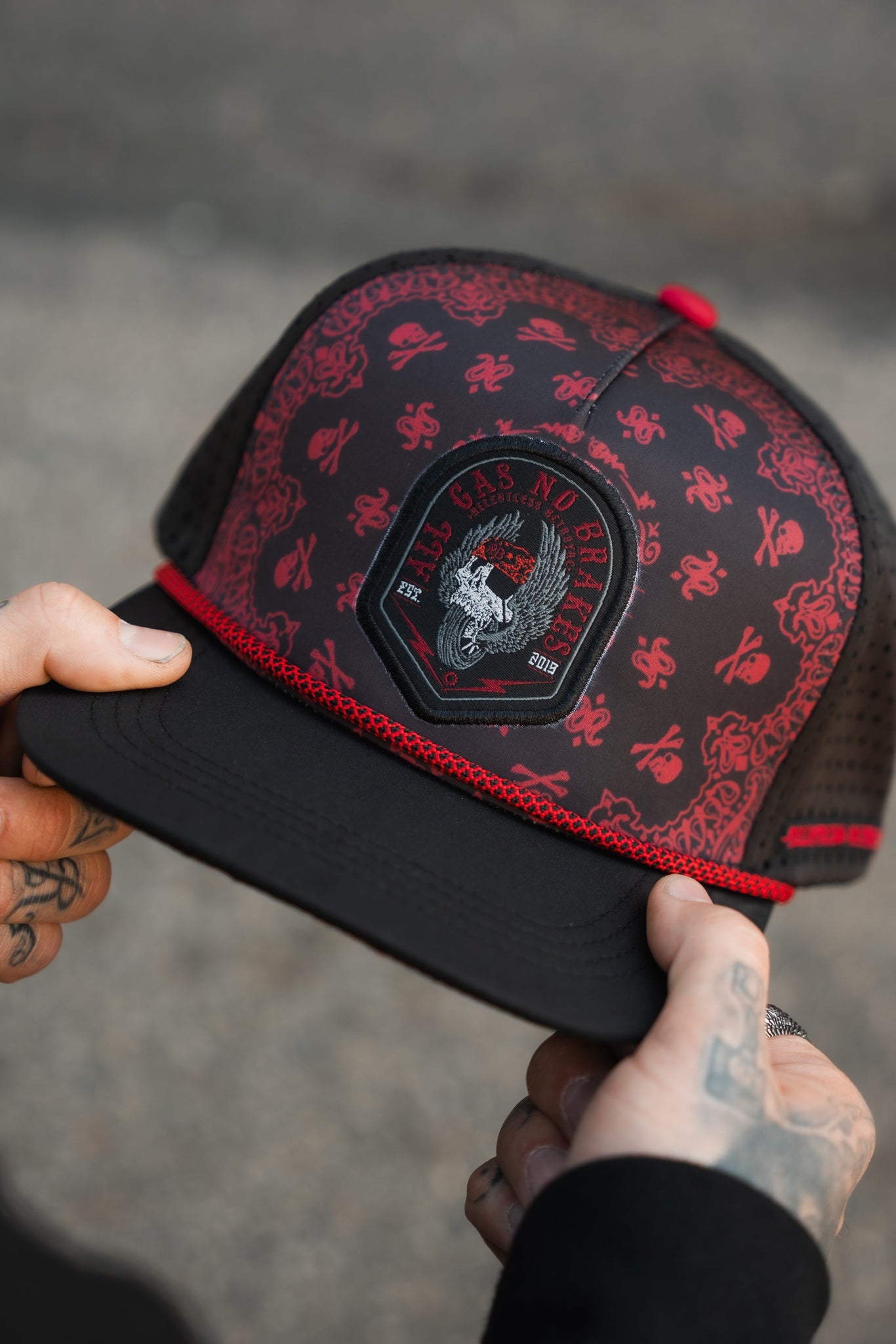 A tattooed person holds the Relentless Betrayal All Gas No Brakes Premium Snapback, a cap featuring skull and crossbones, bandana prints, black mesh sides, a red trim, and the No Gas, No Brakes patch for an edgy look.