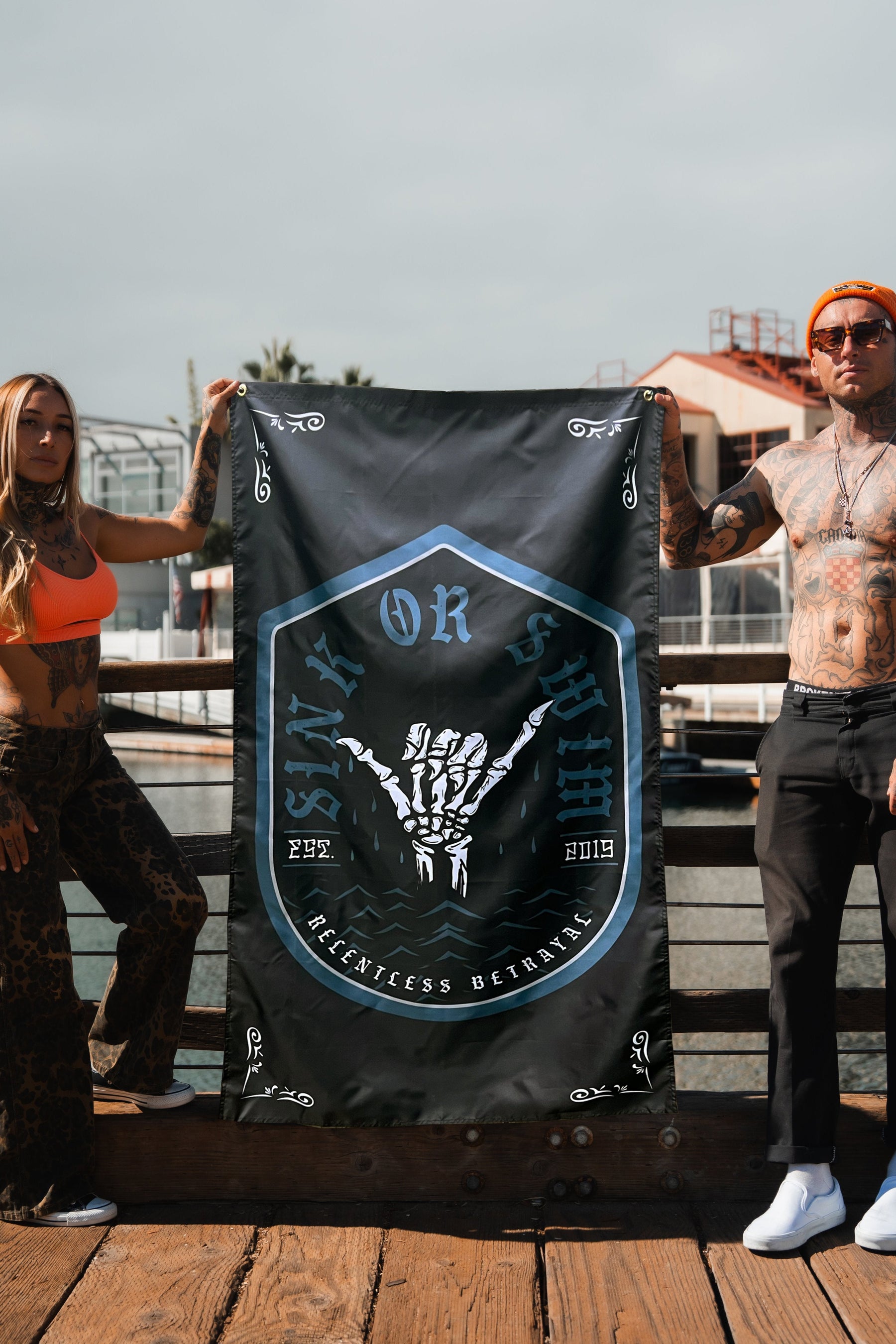On a wooden dock, two individuals hold the Sink or Swim 2.O Flag by Relentless Betrayal, displaying a skeletal hand design with Sink or Swim, 222, and 2019. The tattooed man sports sunglasses while the woman wears a bright top and leopard pants, both symbolizing resilience.
