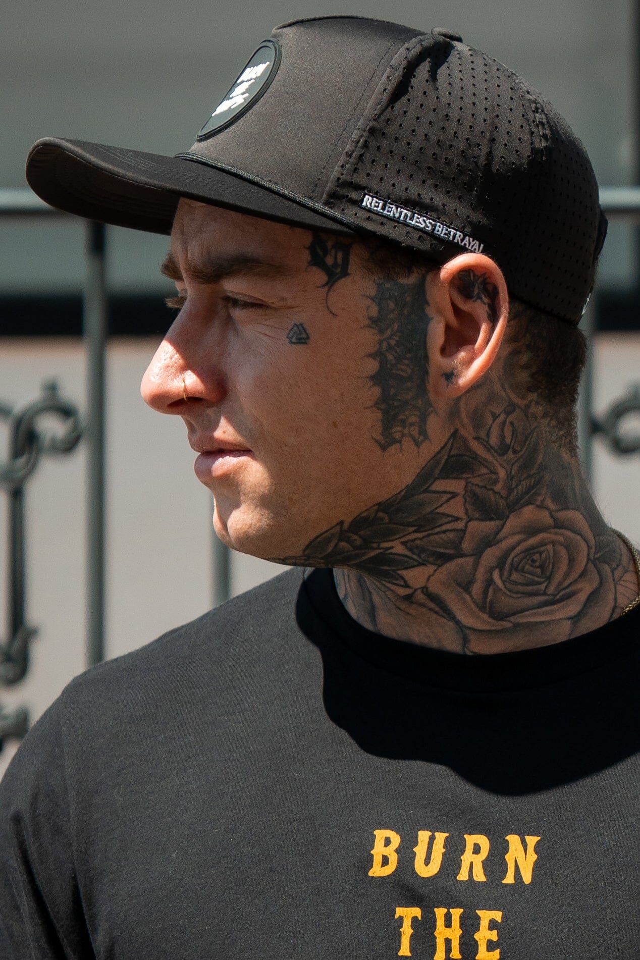 A man in a Burn The Ships Black PVC Snapback from Relentless Betrayal gazes left. His face and neck tattoos, featuring a rose and teardrop, stand out. Hes in a black T-shirt with partially visible yellow text, set against a blurred background hinting at piracy.