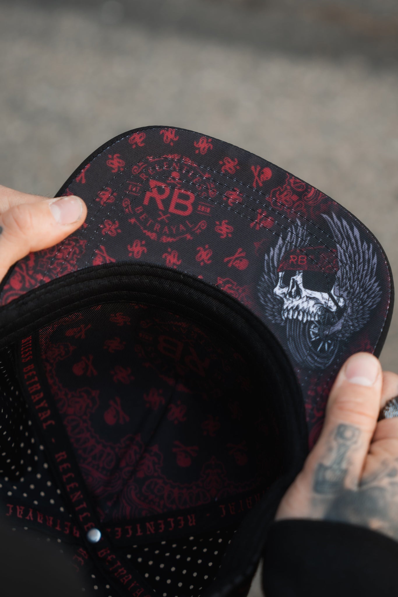 A person grips the All Gas No Brakes Premium Snapback cap by Relentless Betrayal, featuring a red and black skull and crossbones design. Inside are bandana prints with a winged skull and RB initials. The outer rim displays Relentless, while tattooed fingers hold the cap.