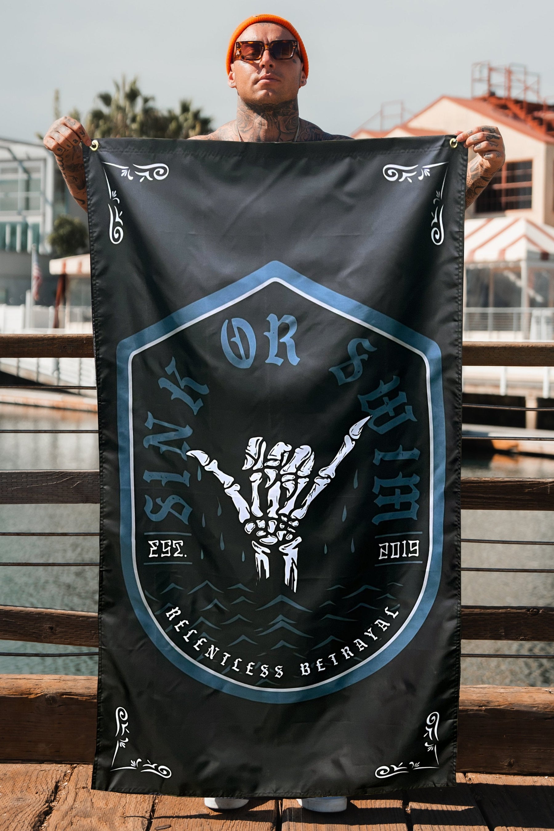 A resilient person stands firm on a wooden platform by water and buildings, holding the black Sink or Swim 2.0 Flag featuring a skeletal hand symbol. They wear an orange beanie and sunglasses, embodying the spirit of the Relentless Betrayal brand.