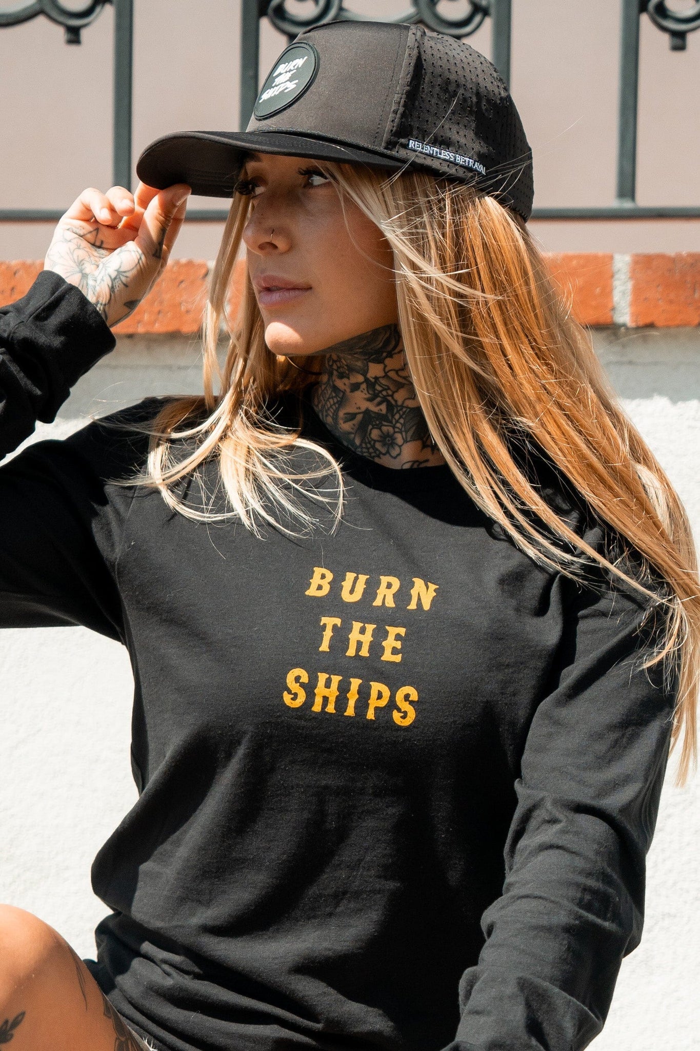 Adorning long hair and tattoos, a person embodies the pirate lifestyle wearing a Relentless Betrayal black shirt with BURN THE SHIPS in yellow and the Burn The Ships Black PVC Snapback. They stand outdoors against a white wall lined with decorative metal railing.