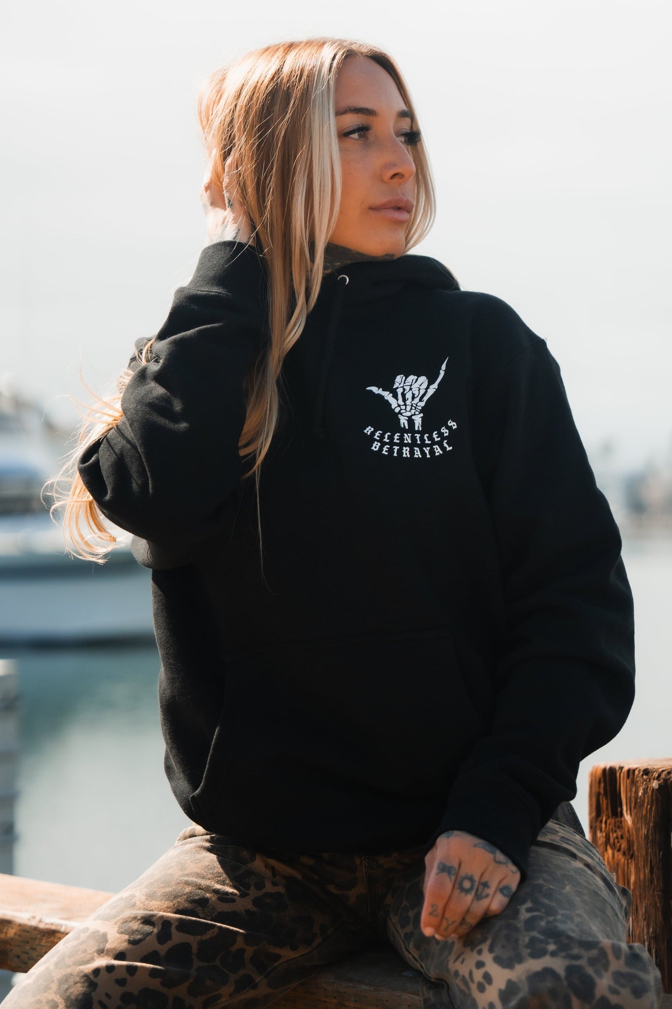 A person with long blonde hair sits outdoors wearing a Relentless Betrayals Sink or Swim Premium Hoodie, embodying resilience. They casually rest one hand on their head, gazing into the distance against a blurred backdrop of water and a boat.