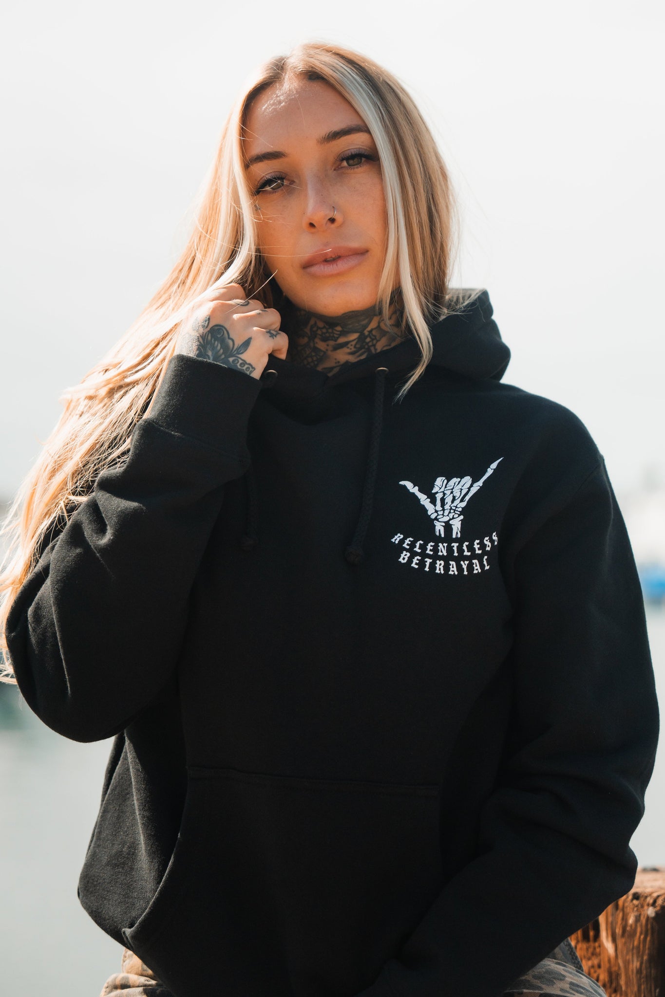 A person with long blonde hair wearing a Relentless Betrayals Sink or Swim Premium Hoodie, featuring a skeletal hand design and text, stands outdoors, embodying resilience. Tattoos adorn their hand and neck, set against a blurred blue sky background.
