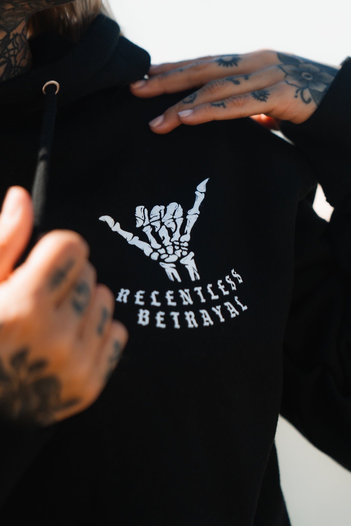 Clad in the Sink or Swim Premium Hoodie by Relentless Betrayal, featuring a skeleton hand rock gesture and RELENTLESS BETRAYAL text, a person with tattooed hands embodies resilience against a vibrant backdrop.