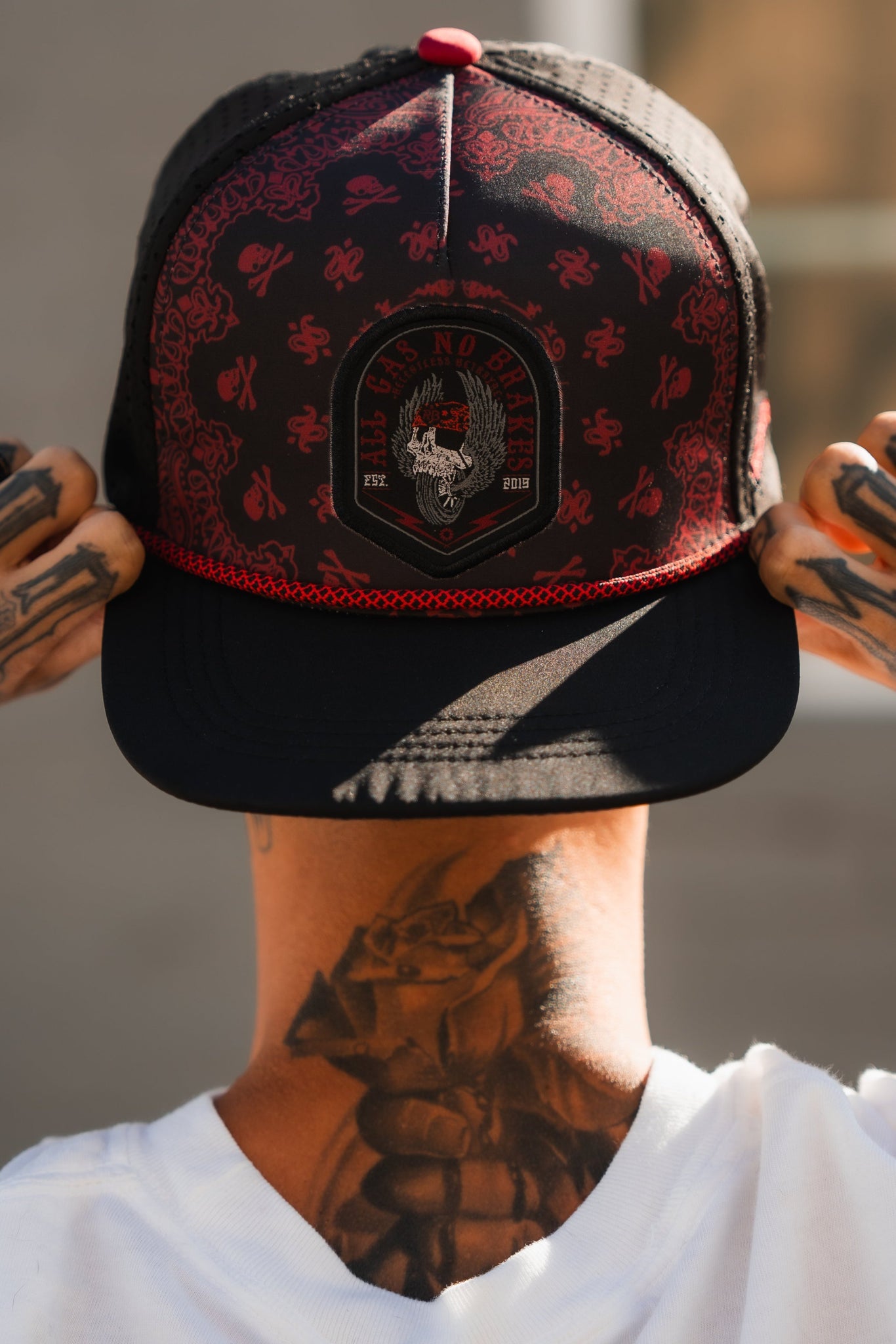 A person sports the Relentless Betrayal All Gas No Brakes Premium Snapback, featuring a black and red dragon bandana print. Tattooed hands hold the brim, while a large tattoo graces their neck. They face away in a white shirt, exuding pure drive.