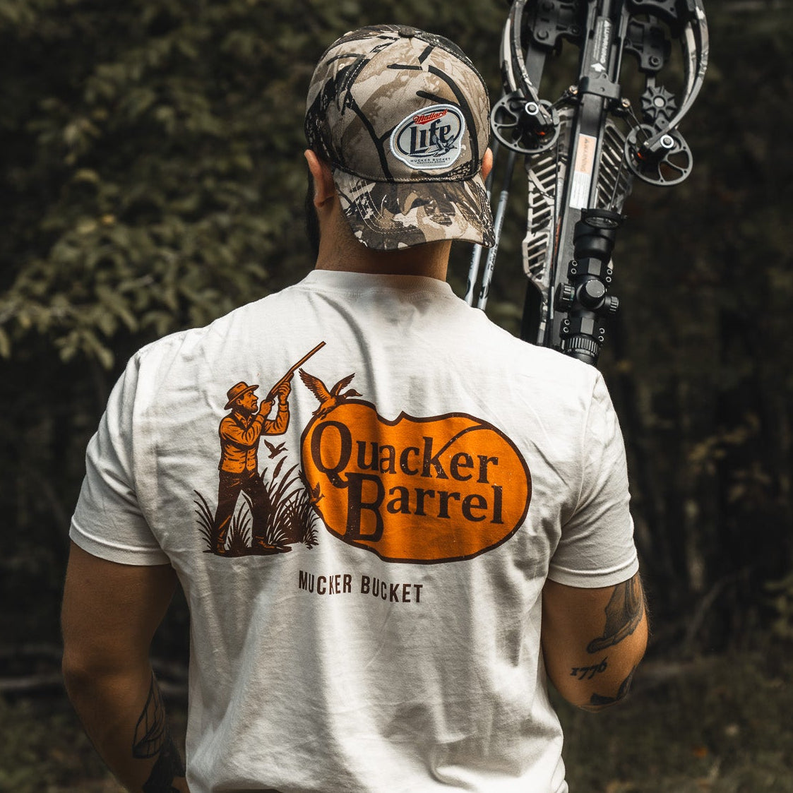 A man in a Mallard Life Deadwood Camo Bucket hat by My Store holds a crossbow over his shoulder. He wears a white shirt with an illustrated hunter and dog and stands in front of trees.