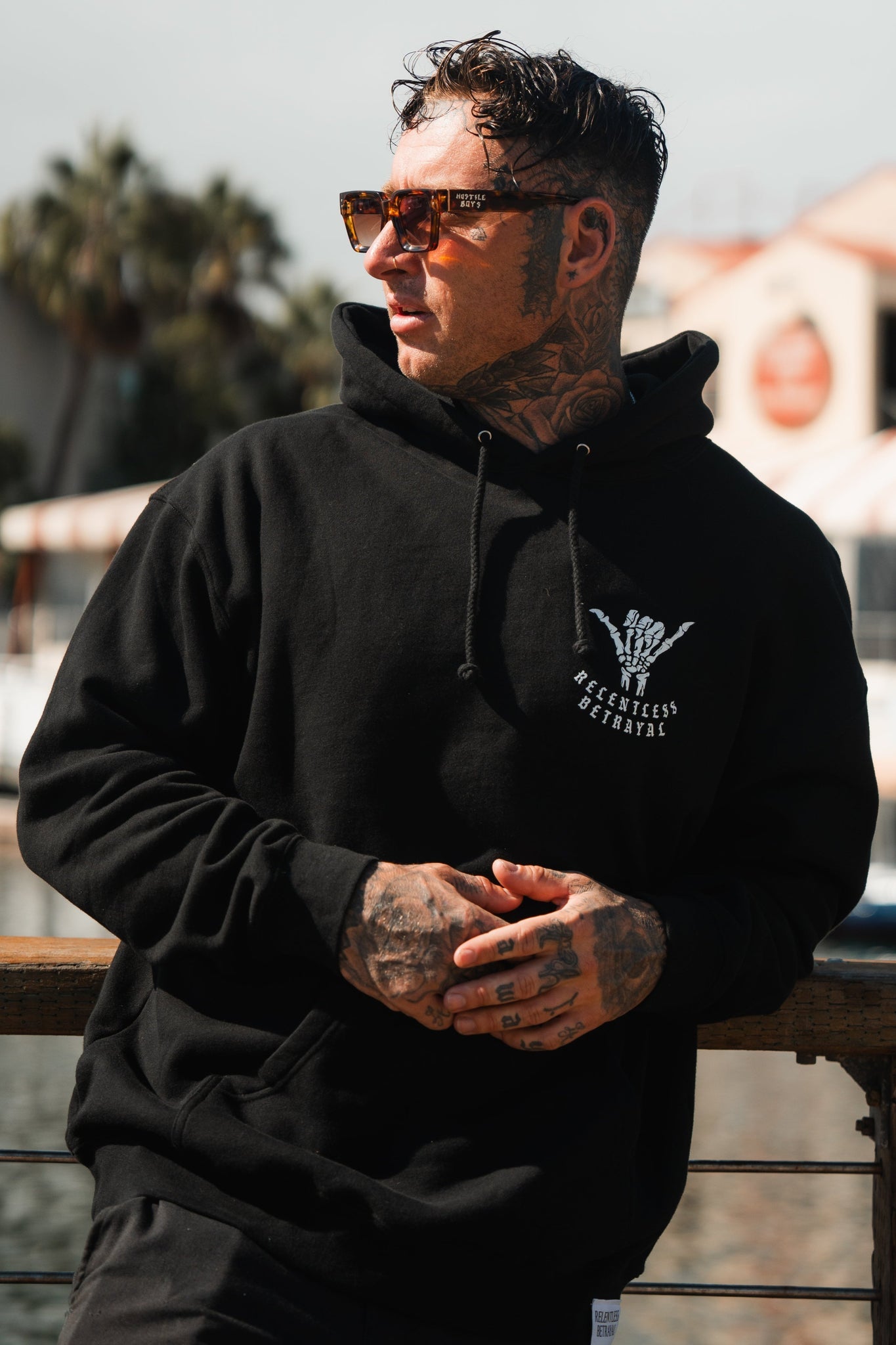 Standing resilient outdoors, a person in a black Sink or Swim Premium Hoodie by Relentless Betrayal pairs with sunglasses, tattoos on hands and neck echoing perseverance. They lean on a railing, framed by palm trees and a building—a testament to overcoming adversity.