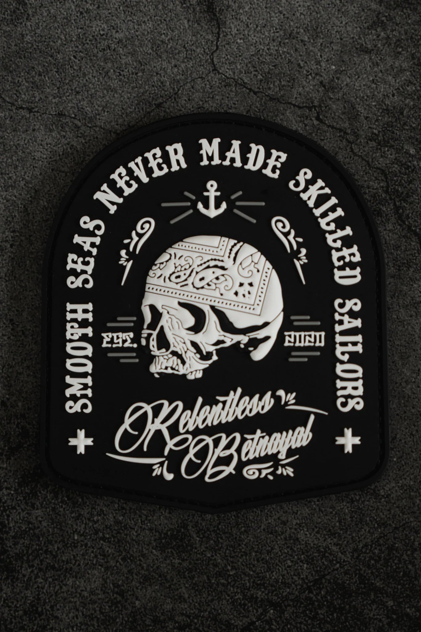 The Skilled Sailor PVC Patch by Relentless Betrayal displays a skull with a bandana, surrounded by an anchor and waves. It features the phrase Smooth Seas Never Made Skilled Sailors and Relentless Betrayal in stylized script on a textured dark backdrop, highlighting the theme of overcoming challenges.