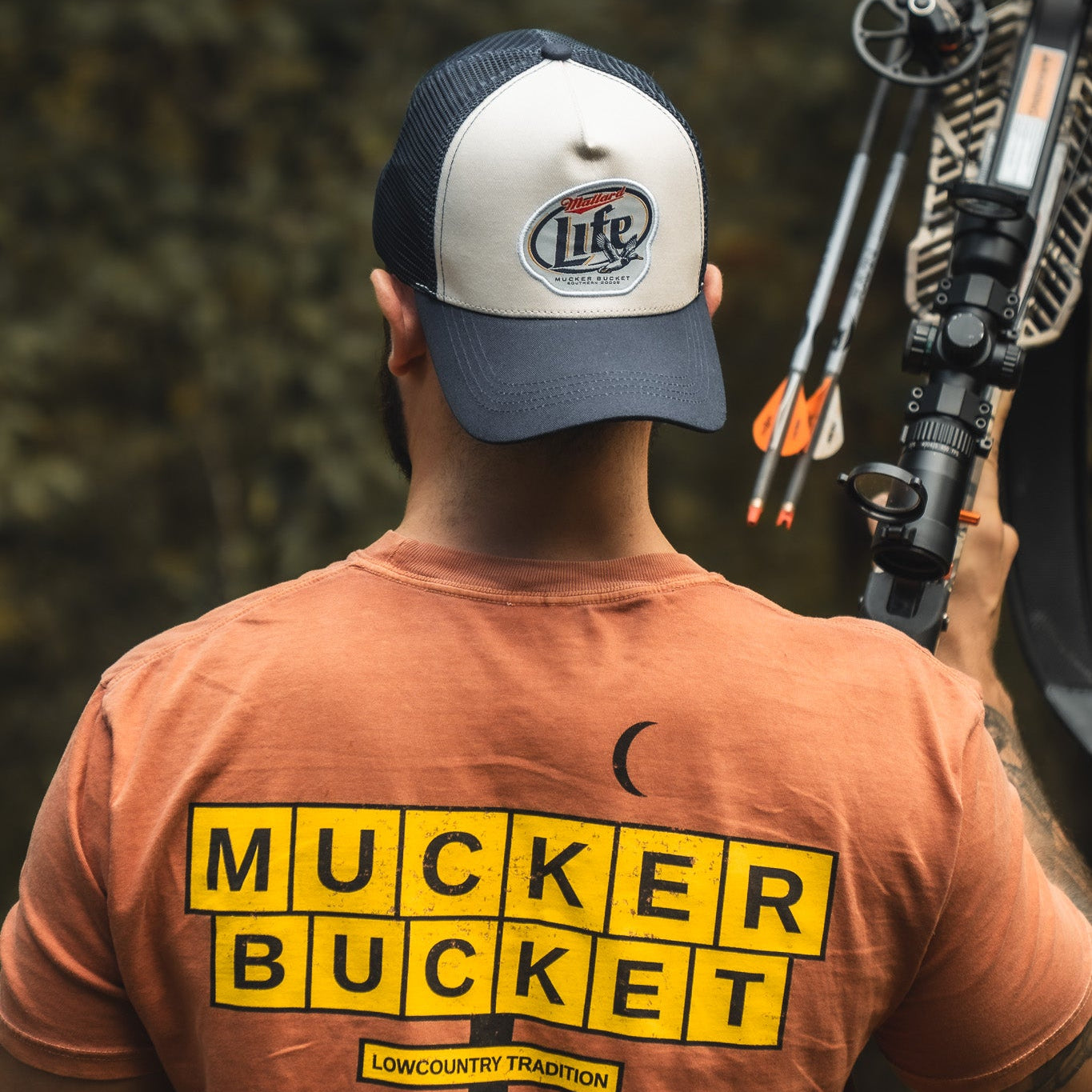 A person wearing a My Store Mallard Life Trucker Bucket hat is seen from behind, holding a compound bow with arrows outdoors.