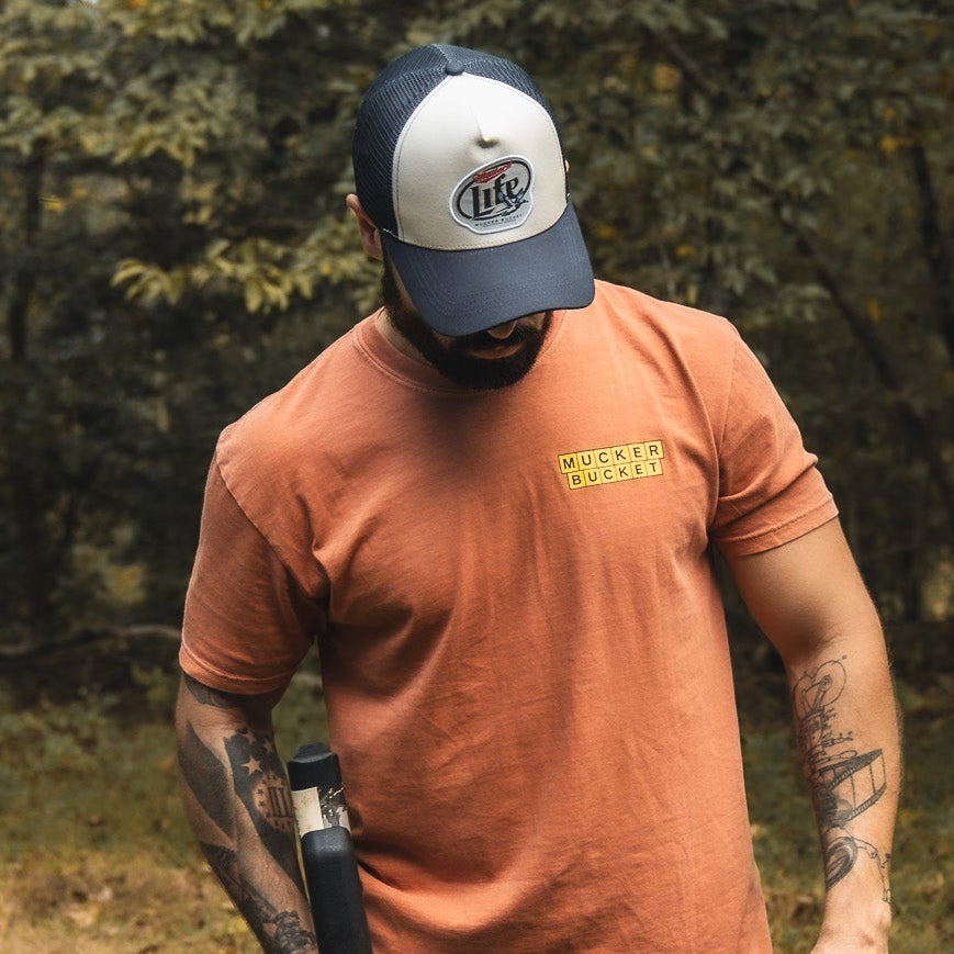 A bearded man with tattoos wearing an orange "Mallard Life Trucker Bucket" T-shirt from My Store and a trucker snapback stands outdoors, looking down while holding a black object, with trees in the background.