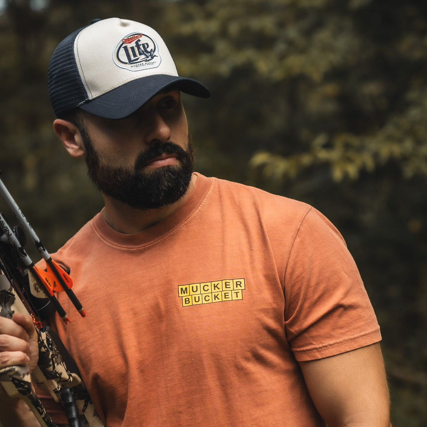 A bearded man wears an orange t-shirt and a black-and-white Mallard Life Trucker Bucket hat by My Store, featuring patch embroidery, while holding a compound bow outdoors with blurred greenery in the background.