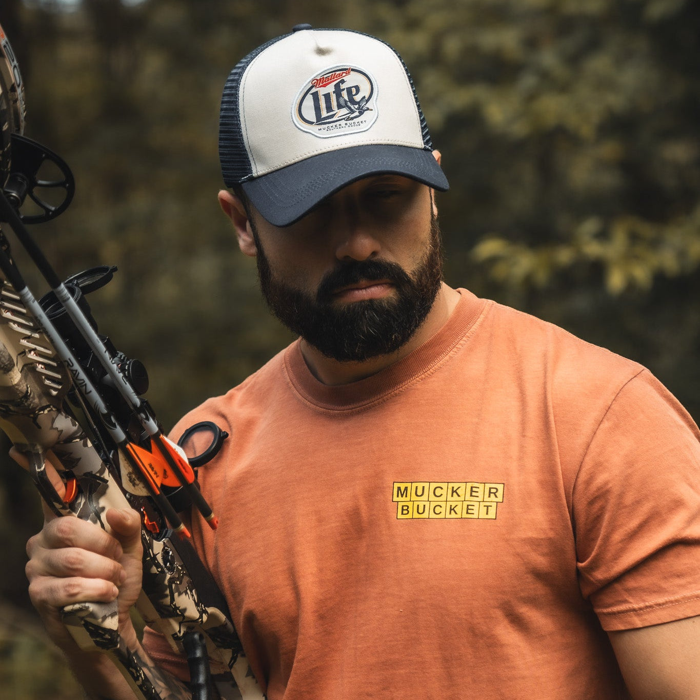 A bearded man in an orange t-shirt and a black-and-white My Store Mallard Life Trucker Bucket hat with patch embroidery holds a camouflage compound bow outdoors, blurry trees in the background.